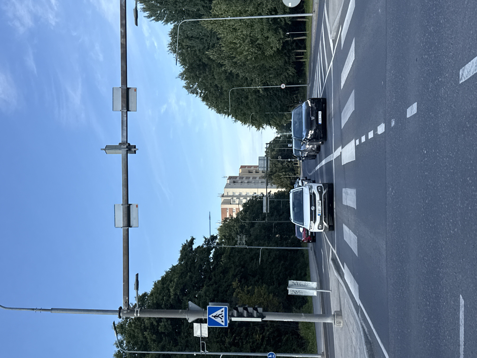 The image depicts a street scene in Tallinn, Estonia, on a clear day with a blue sky and scattered clouds. The perspective is from a vehicle moving forward. The foreground shows a paved road with white pedestrian crossing lines and lane dividers. In the middle ground, several cars are visible. A white Volkswagen SUV is in the right lane, its license plate reading "691 JNP". To its left, a black Peugeot sedan is in the left lane, with license plate "069 TYG". Behind these vehicles, other cars are partially visible. On the left side of the road, a pedestrian crossing sign is mounted on a pole, accompanied by traffic lights. Further down the road, trees line both sides, with a tall residential building visible in the distance beyond the trees on the left. Streetlights are also present along the road. The overall scene suggests a typical urban street during daylight hours.