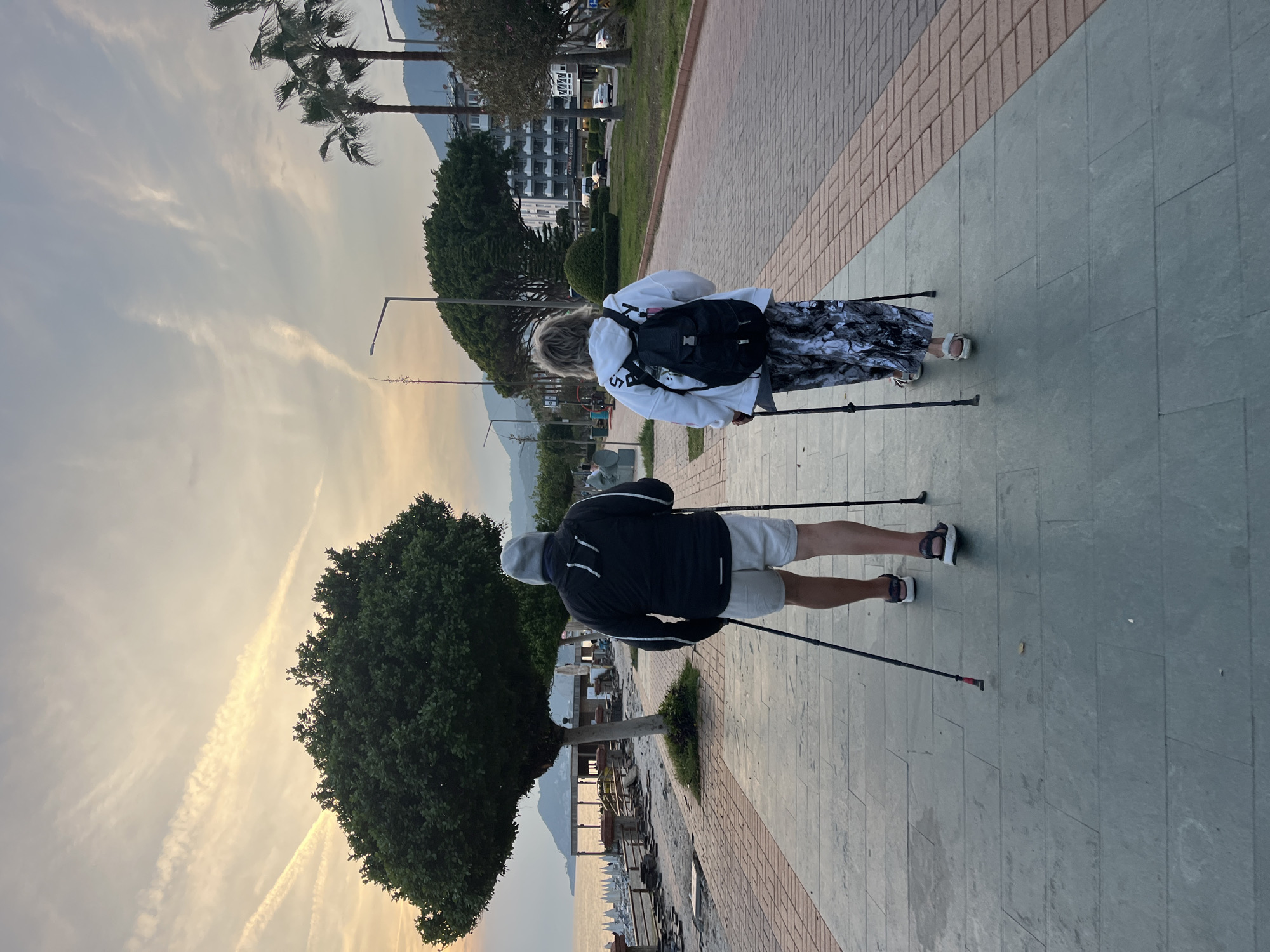 Two individuals are walking side-by-side along a paved walkway in Alanya, Turkey. The first person is a man wearing a dark jacket, light-colored shorts, a gray hat, and sandals. He is using walking poles. The second person is a woman with blond hair wearing a white sweatshirt, patterned pants, sandals, and a black backpack. She is also using walking poles.
The walkway is paved with a combination of gray stone and brick. The scene is outdoors, with trees and other greenery visible along the walkway. Buildings and a mountain are visible in the distance. The sky has a hazy appearance, likely due to the sunset or sunrise. A body of water with boats is visible near the left side of the image.