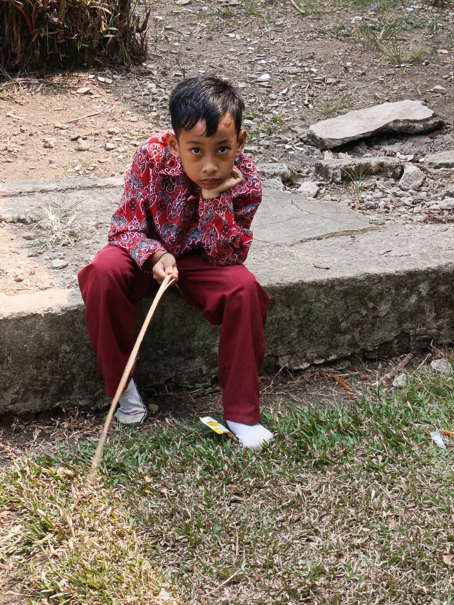 A young boy, appearing to be of Southeast Asian descent and approximately 6-8 years old, sits on a concrete ledge. He is wearing a vibrant red shirt with a repeating pattern of white and blue motifs and dark red pants. His left hand rests on his chin, and his right hand holds a thin, light-brown stick, which he is using to poke at the grassy ground in front of him. He is looking directly at the camera with a neutral expression. The background consists of dry, gravelly earth with scattered rocks and dry vegetation, and a patch of green grass in the foreground. The scene suggests an outdoor setting, possibly in a park or a less-maintained area, within the city of Megang Sakti IV, Indonesia, given the context.