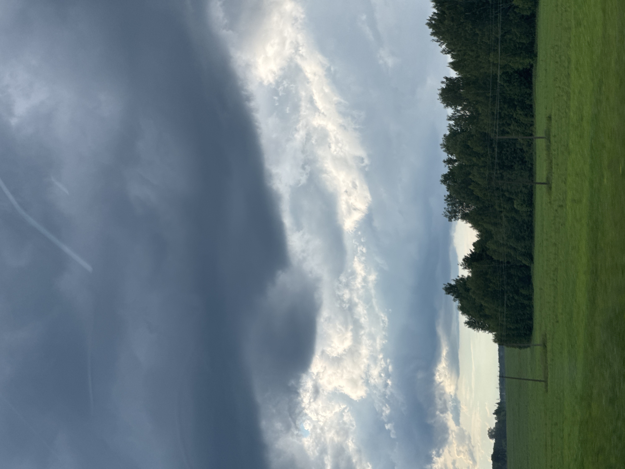 A wide-angle, eye-level shot captures a rural landscape under a dramatic sky. The foreground is dominated by a vast, flat, green field, suggesting it is either a pasture or a cultivated area. Two sparsely constructed, rustic wooden poles with wires stretched between them stand in the field, indicating some form of agricultural or utility infrastructure.
In the middle ground, a dense line of dark green trees forms a distinct barrier across the horizon, with some lighter green foliage visible on the right side of the tree line. The background is entirely consumed by a turbulent sky filled with layered clouds. The upper portion of the sky is a dark, ominous gray, transitioning to lighter grays and whites in the middle, and finally to a brighter, almost luminous band of white clouds towards the bottom, with hints of blue sky peeking through. Faint white contrails from aircraft are visible in the upper sky. The overall lighting suggests either an approaching storm or the aftermath of one, with diffused light illuminating the scene.
The location is identified as Krasnoyarsk, Russia, implying a natural or semi-natural environment within or near the city. There are no people or animals visible in the image, nor are there any discernible structures beyond the fence posts and the distant tree line. The scene is static and devoid of any human activity. The image appears to have been taken from a moving vehicle, as there is a slight motion blur on the grass in the immediate foreground, particularly on the left side.