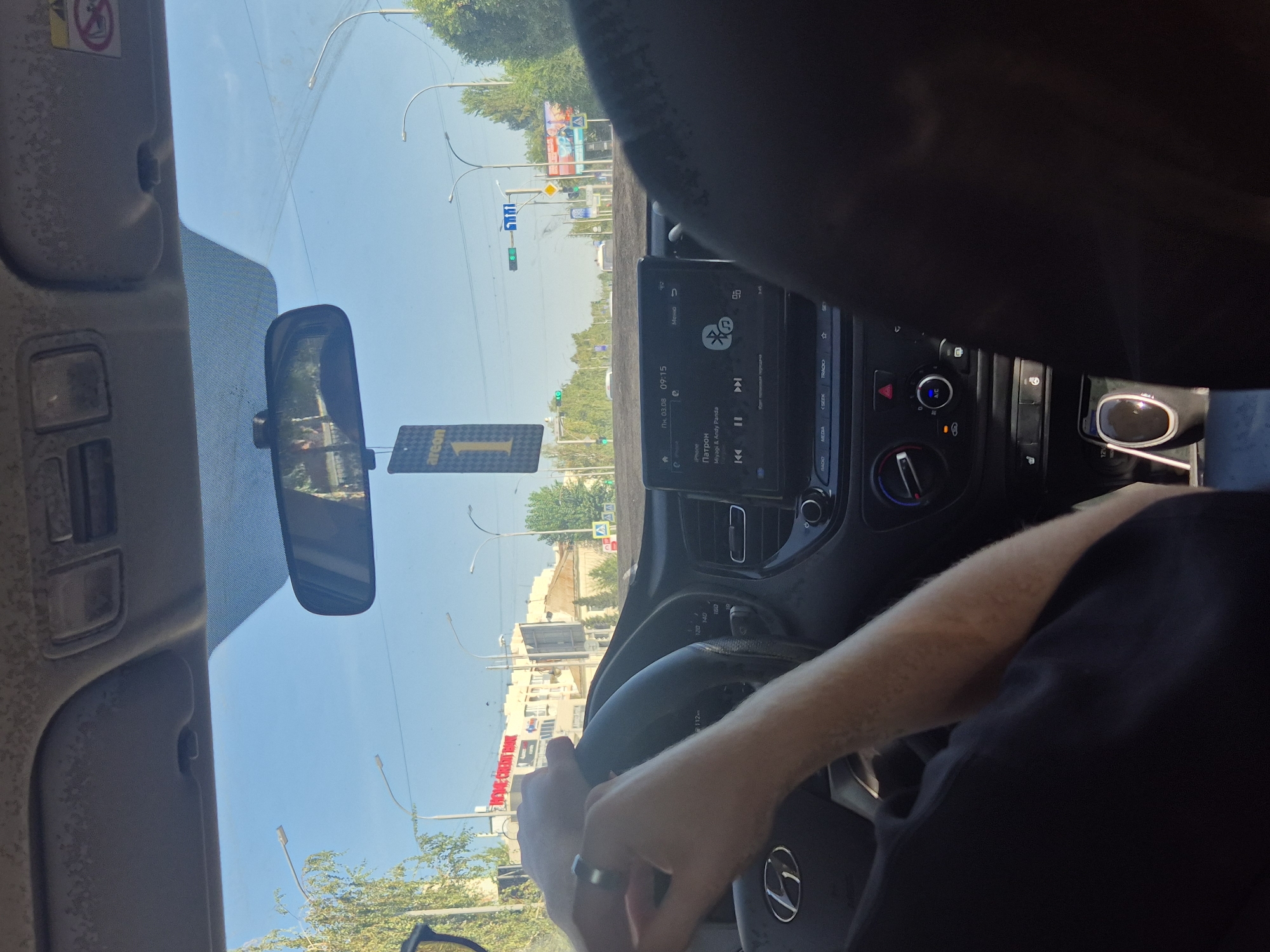 The image captures the interior of a car, viewed from the passenger seat, looking forward. The driver's hands are visible on the steering wheel, one hand with a ring. A car air freshener, branded "areon 1," hangs from the rearview mirror. The car's dashboard features a touch screen displaying music information, showing the song "Паттрон" by Mirage & Andy Panda, and the time 09:15. Traffic lights and streetlights are visible through the windshield, along with buildings and trees lining a street. A sign for "HOME CREDIT BANK" is clearly discernible on a building in the distance. The scene is set during the daytime, with a clear blue sky overhead. This image is consistent with a view from a vehicle on a street in Kokshetau, Kazakhstan, given the architectural styles and signage.