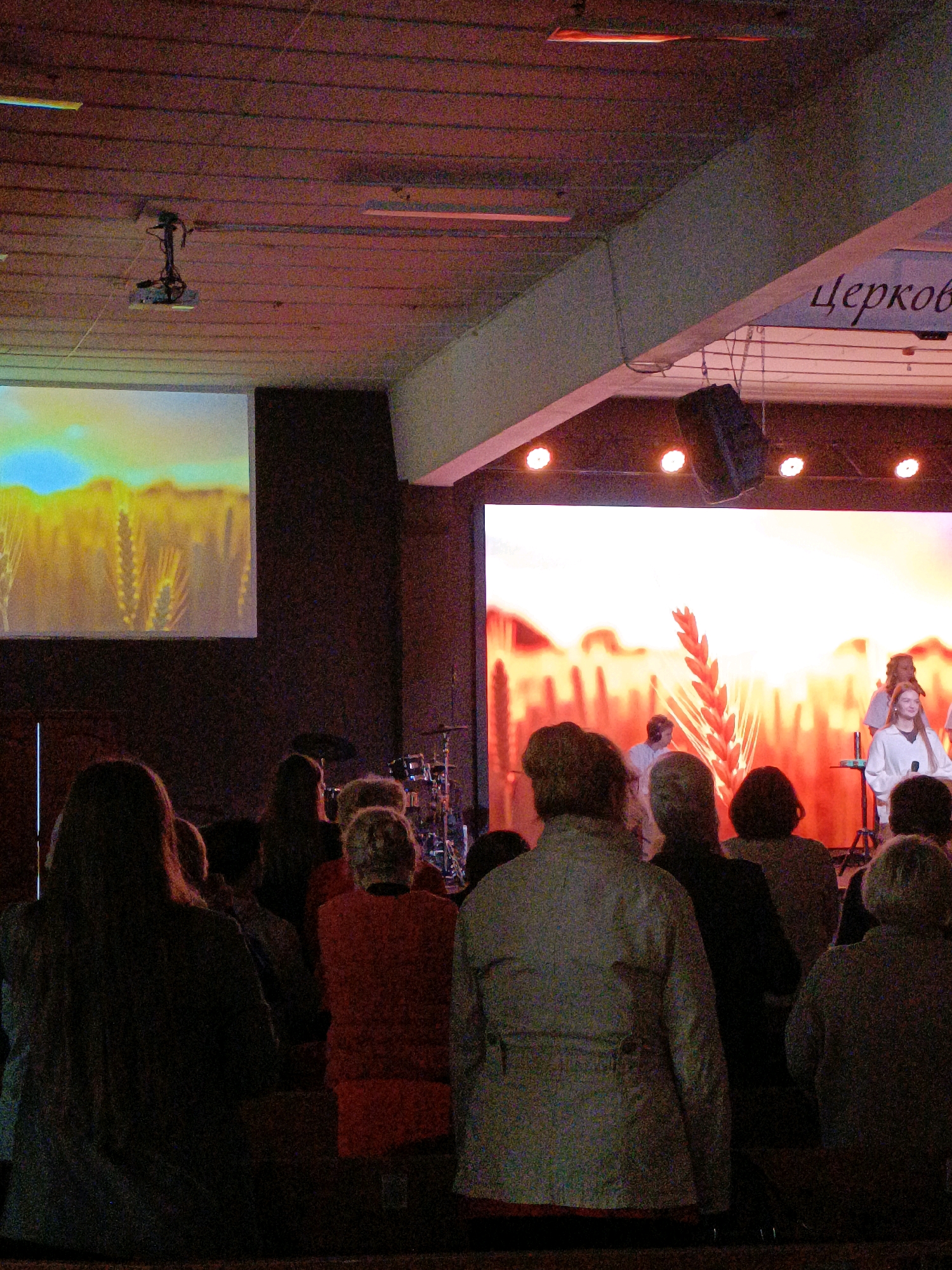 The image depicts an indoor gathering, likely a religious service, held in Asbest, Russia. In the foreground, a crowd of individuals are seated, facing a large projection screen. The screen displays a digitally rendered image of a golden wheat field against a bright sky. To the left, another screen shows a similar pastoral scene. A stage area is visible in the background, featuring a group of people, some appearing to be performers, with musical instruments and microphones present. The overall lighting is dim, with stage lights illuminating the performers and creating a warm glow. The architecture suggests a modern community hall or church interior, with exposed wooden ceiling beams and overhead lighting fixtures. A projector is mounted on the ceiling. The text "Церков" (Church) is visible in Cyrillic script on a banner overhead.