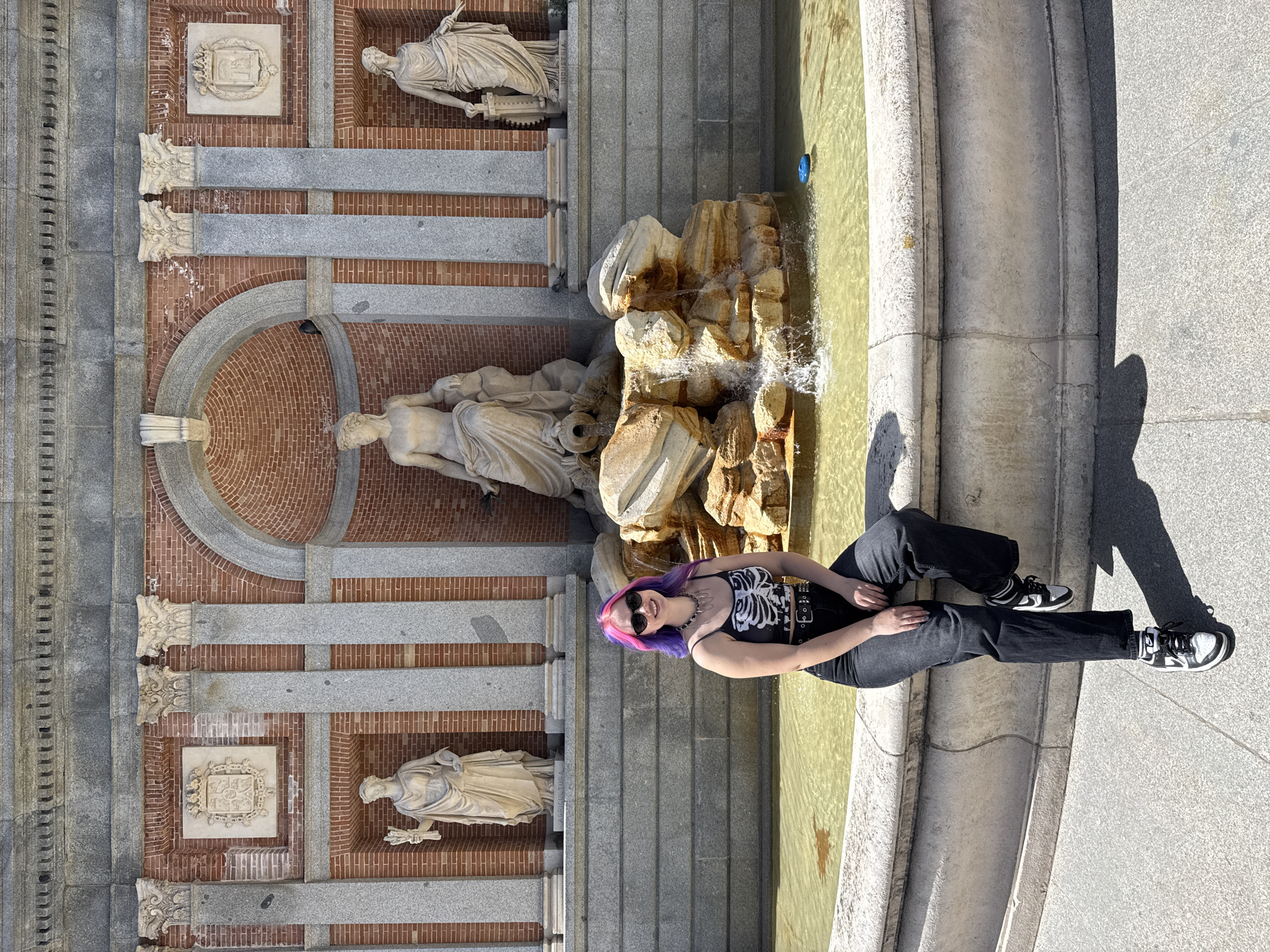 A young woman with purple and pink hair is sitting on the edge of a fountain in Madrid, Spain. The fountain is located in front of a brick wall with stone pillars and statues. The woman is wearing a black tank top, black pants, and black and white sneakers. She has sunglasses on and is looking at the camera. The water in the fountain is a greenish-yellow color. The ground around the fountain is made of stone. The statues are facing to the right.