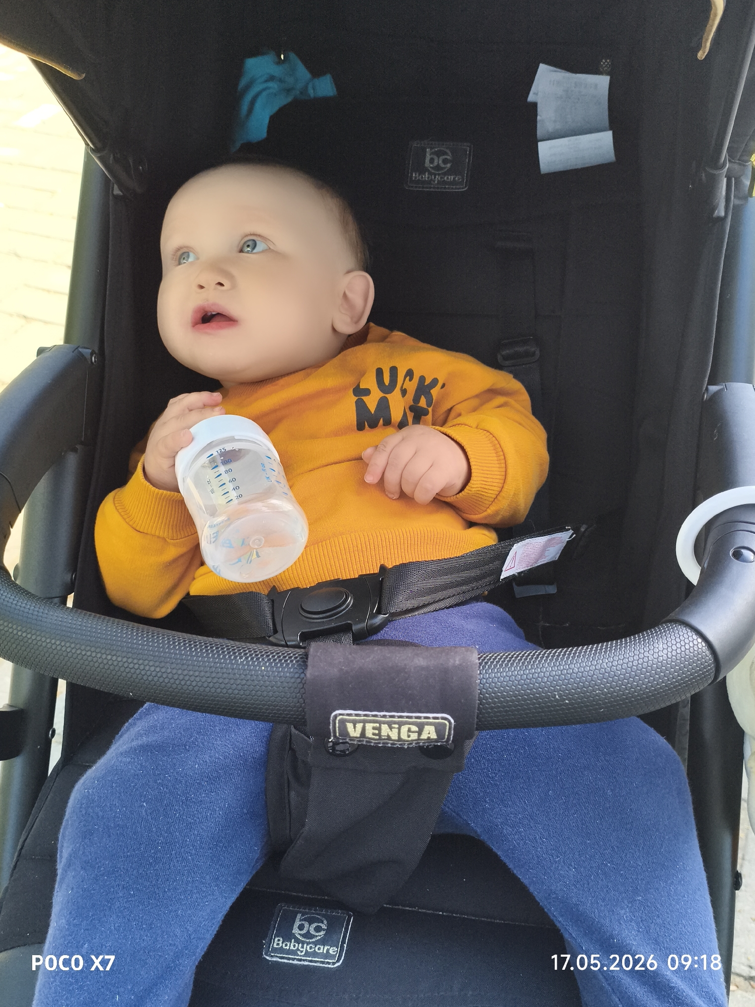 A baby with blue eyes and blonde hair sits in a black stroller, holding a feeding bottle in their left hand. The baby wears a mustard-yellow sweater with the words "LUCK MAN" printed on it and blue pants. The stroller has a black safety bar and a Venga brand tag. The Babycare brand is seen in two places on the stroller. The date "17.05.2026 09:18" is imprinted in the lower right corner, and "POCO X7" is in the lower left corner. The location is Chaikovsky, Russia.