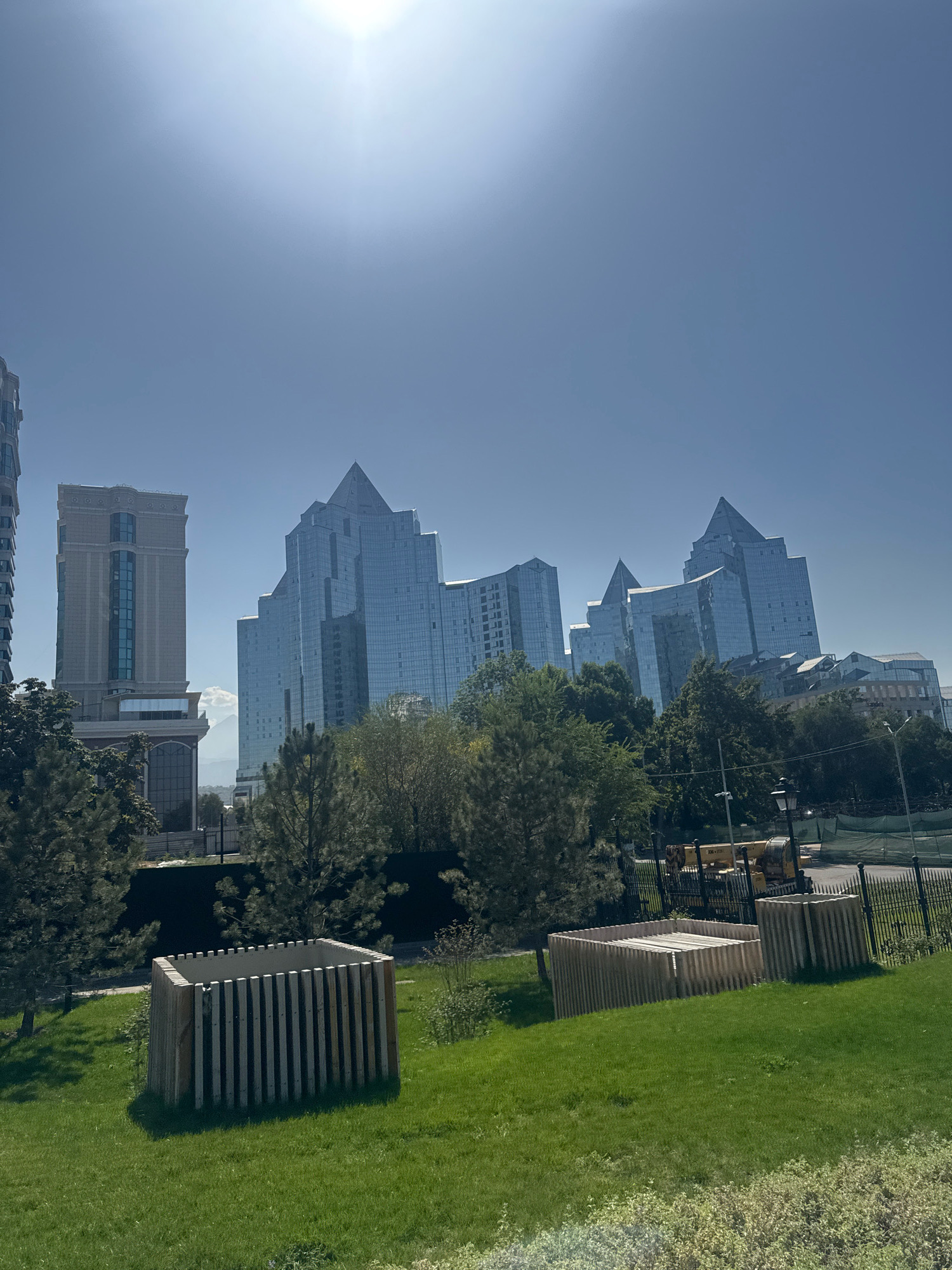 This is a daytime, eye-level, wide-angle photograph of a modern urban park area with a city skyline in the background. The sun is high in the sky and is partially obscured by a lens flare in the upper center of the image. The foreground features a gently sloping, well-maintained lawn with two wooden, slatted structures, possibly planters or seating areas, on the grass. A black metal fence runs along the edge of the park, separating it from a paved area beyond. In the mid-ground, there are several mature trees and some construction equipment, including a yellow crane. Behind the trees, a cluster of modern glass-fronted skyscrapers dominates the background. The building on the left is lighter in color with a more traditional architectural style, while the buildings to the right are characterized by sharp, angular designs and reflective glass facades, giving them a futuristic appearance. The sky is a clear, light blue. The scene is located in the city of Almaty, Kazakhstan, likely in the vicinity of the "Baiterek" monument area or a similar central business district known for its modern architecture.