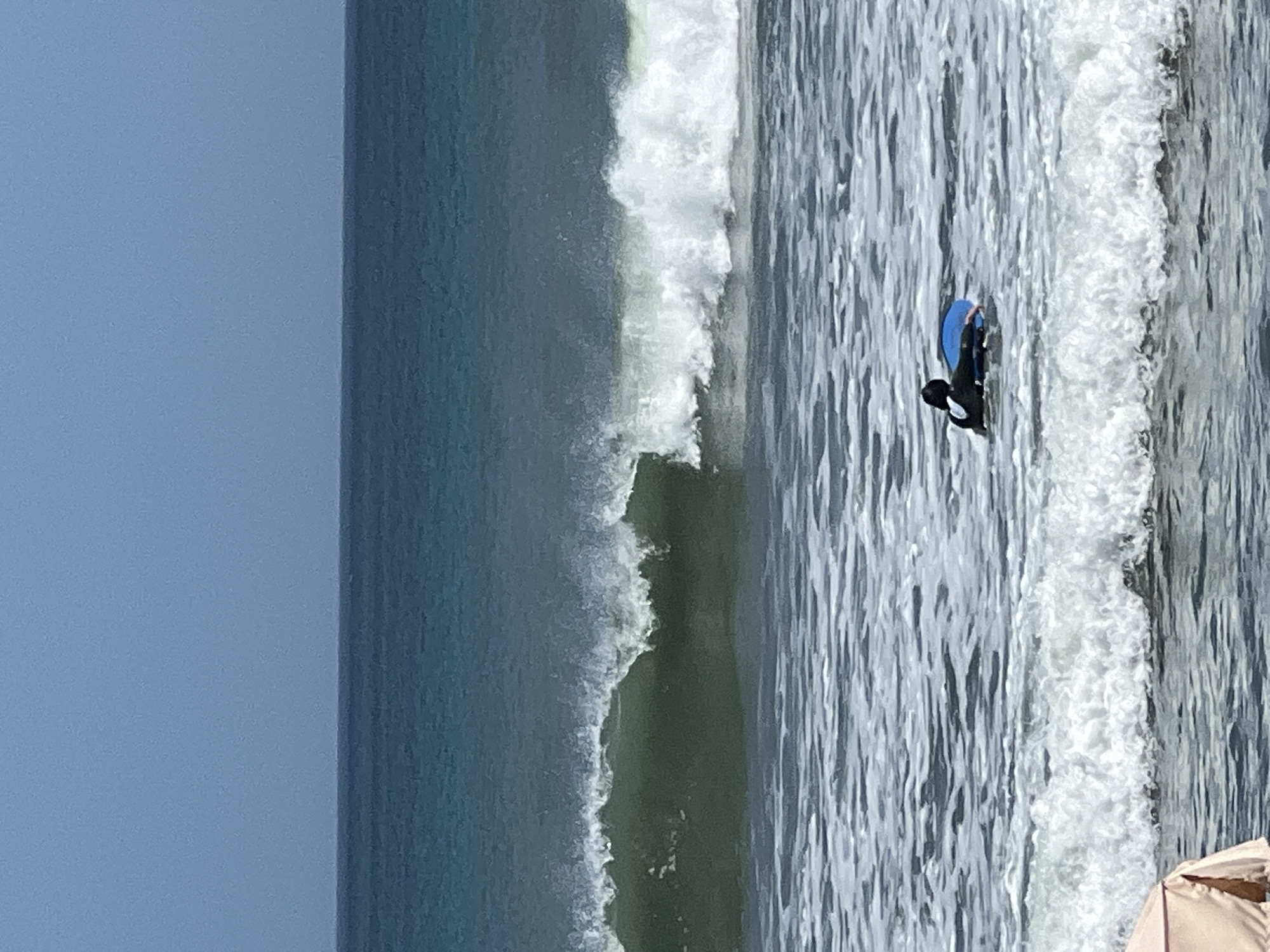 In the ocean off Kuta, Indonesia, a person wearing a black wetsuit and a white rashguard is lying on a blue bodyboard. The person is facing towards the left of the frame and appears to be paddling. The ocean is choppy with whitecaps, and a large wave is breaking in the middle ground of the image. The sky is clear and blue. In the foreground, a portion of a tan beach umbrella is visible.