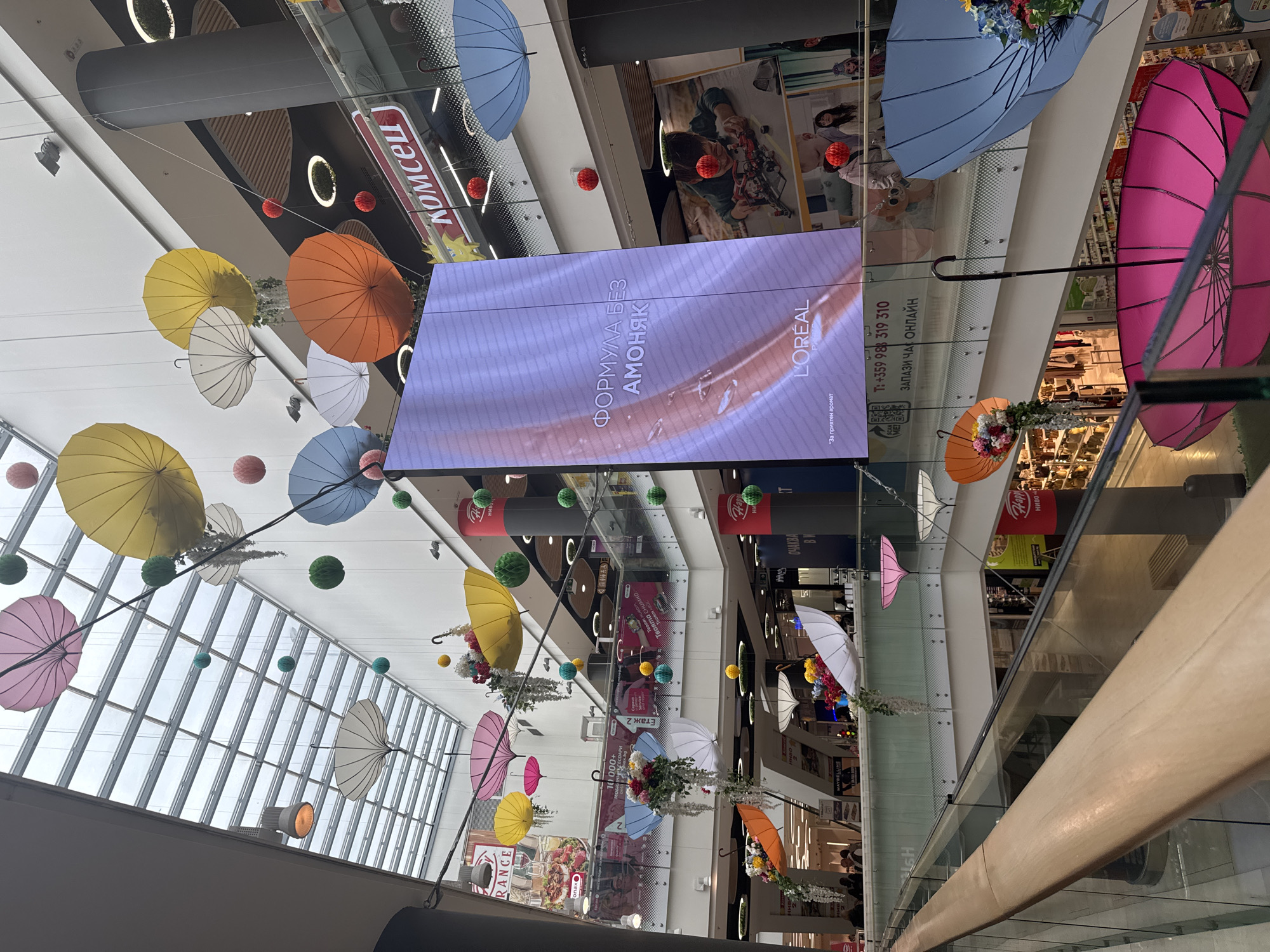 This upward-angled view captures the vibrant interior of a multi-story shopping mall in Sofia, Bulgaria. The central focus is on a large digital screen displaying an advertisement for L'Oréal hair product, featuring the Cyrillic text "ФОРМУЛА БЕЗ АМОНЯК" (FORMULA WITHOUT AMMONIA) and "*За приятен аромат" (*For a pleasant aroma).
The mall's ceiling and upper levels are adorned with numerous colorful open umbrellas in shades of yellow, orange, white, pink, and blue, many accented with artificial flowers and greenery. Spherical decorations in green, red, and yellow also hang at various heights, contributing to a festive atmosphere. A large glass skylight structure is visible on the left, letting in natural light.
The architecture showcases multiple levels with glass railings and light-colored surfaces. In the foreground, a wooden handrail marks the viewer's position on an upper floor. Below, glimpses of different shops and stores are visible, some with promotional posters. One sign clearly displays a phone number "+359 983 319 310" and "ЗАПАЗИ ЧАС ОНЛАЙН" (BOOK AN APPOINTMENT ONLINE), alongside a QR code. Other partially visible Cyrillic text includes "Комсец" (Komsets, possibly a store name), "Етаж 2" (Floor 2), "Ниво" (Level), "ОЧАКВА" (EXPECTS), and "Твоята снимка" (Your photo). A few blurry figures of people can be seen on the lower levels, engaged in typical mall activities. The overall impression is bright, inviting, and well-decorated, likely enhancing the shopping experience.