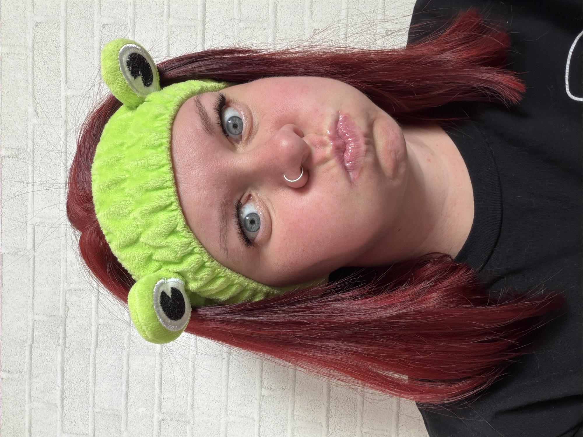 A portrait-style image, taken in Chaikovsky, Russia, depicts a person with bright red, medium-length hair. The individual is wearing a bright green, fuzzy headband featuring two prominent frog eyes positioned on top. The subject's face exhibits wide-open blue eyes, a flushed complexion, puffed cheeks, and pursed, glossy lips, conveying a playful or surprised expression. A silver-colored hoop ring is visible in the right nostril. A black, round-neck t-shirt is partially visible on the subject's torso. The background consists of a uniformly white brick wall with visible grout lines.