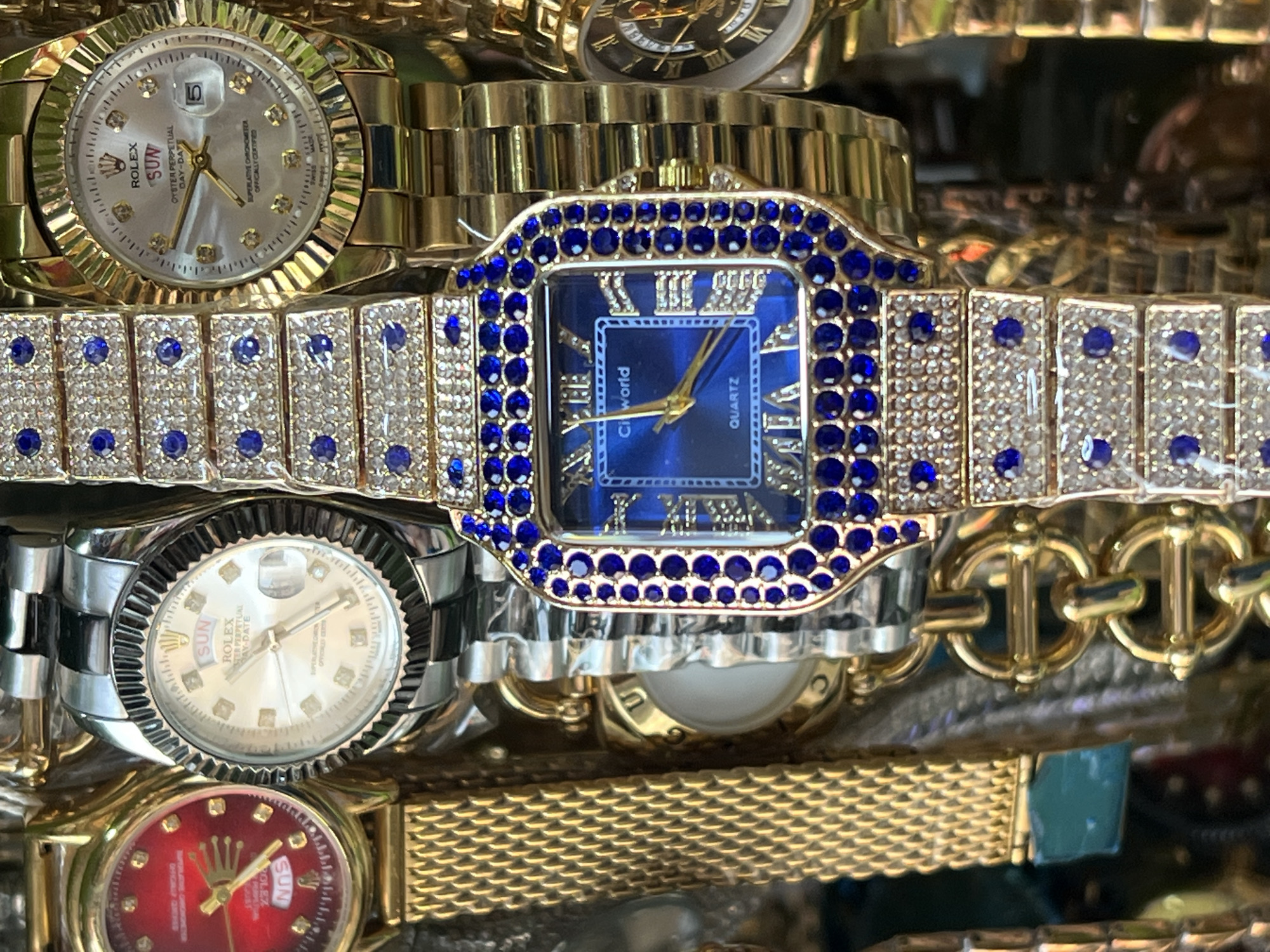 This is a close-up, eye-level, still-life photograph of a collection of luxury wristwatches. The watches are gold-toned and appear to be either real or high-quality replicas, featuring various designs and embellishments. One prominent watch in the foreground has a square, blue face surrounded by a band encrusted with clear rhinestones and blue gemstones. To its left, a watch with a silver band and a white face, possibly a Rolex, is visible. Another watch on the far left has a red face and a gold mesh band. Other watches with gold bands and various face colors are also visible, suggesting a display or collection of timepieces. The background is a blur of metallic and colored surfaces, indicating that the watches are likely displayed together, perhaps in a store or market stall. The lighting is bright and even, highlighting the intricate details and reflective surfaces of the watches. The image does not contain any people or any indications of the city of Bukuru, Nigeria.