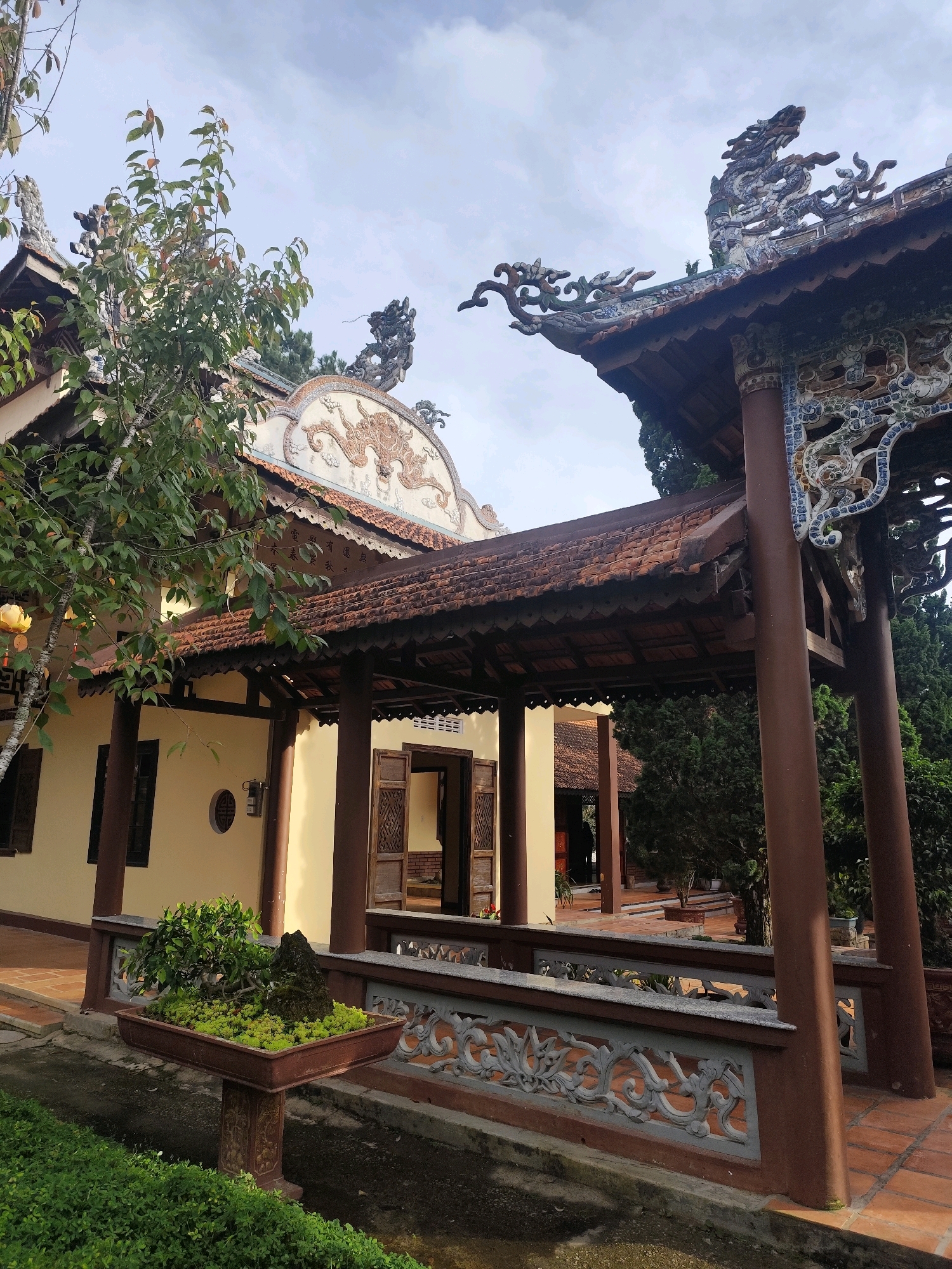The image shows a close-up view of a portion of a Buddhist temple in Da Lat, Vietnam. The temple features traditional Vietnamese architecture with ornate dragon motifs on the roof and decorative eaves. A covered walkway with a tiled roof supported by brown pillars leads into the building. In the foreground, a beautifully crafted bonsai tree sits in a rectangular pot on a decorative stand. The surrounding area includes lush green foliage and a paved courtyard. The sky is overcast with scattered clouds.