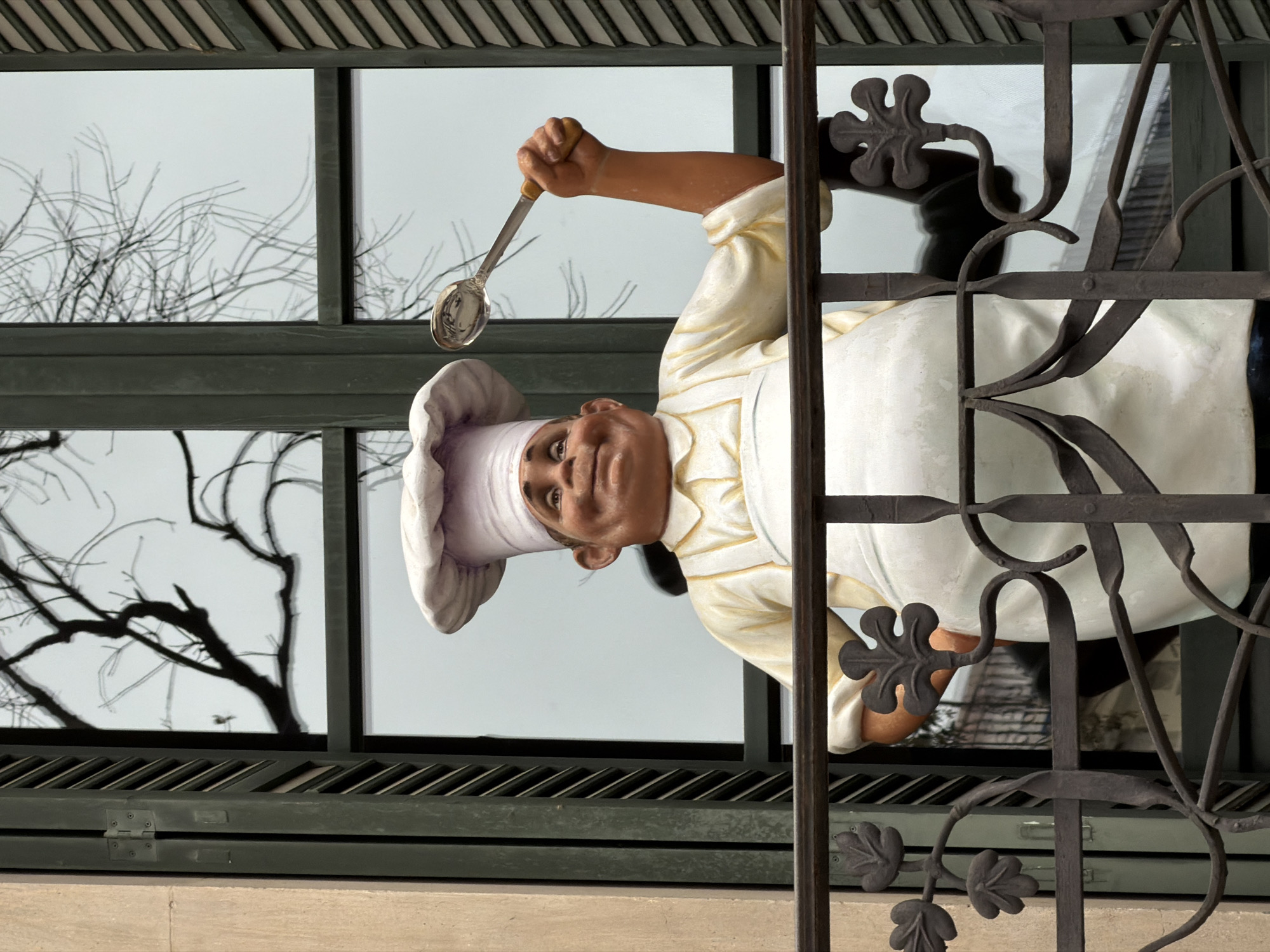 An outdoor decoration featuring a cheerful chef figure is depicted, rotated 90 degrees counter-clockwise, appearing to hang upside down from a building facade in Barcelona, Spain. The statue portrays a male chef from the waist up, with a ruddy complexion, a pleasant smile, and bright, upturned eyes. He wears a classic white chef's hat (toque) and a light-colored, possibly off-white or cream, chef's jacket. His right arm is extended, holding a silver ladle with a golden handle, which reflects light.
The chef figure is positioned behind an ornate, dark metal grille or railing, characterized by scrolling patterns and leaf-like embellishments that partially obscure the view. Behind the chef, a large window reveals a bright, overcast sky and the intricate network of bare tree branches, suggesting a winter or early spring setting. Below the window, a dark green horizontal structure with slatted details, possibly part of a shutter or ventilation system, is visible, resting on a beige stone ledge. The lighting is diffused, indicating a bright day without direct sunlight. There is no visible text in the image.