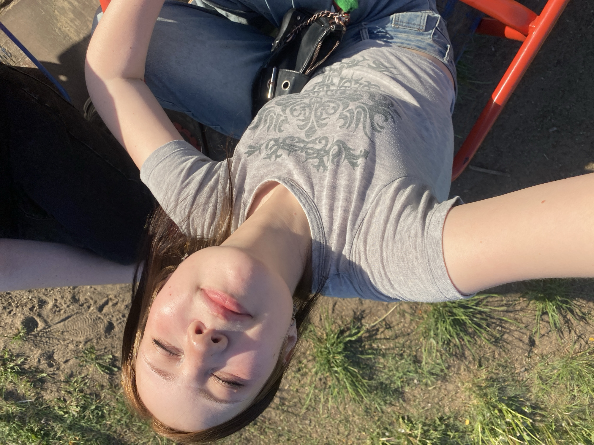 A young woman is lying on her back on the ground, facing upwards towards the camera. She has fair skin, closed eyes, and light pink lips. Her hair is brown with lighter highlights and is parted to the side. She is wearing a gray t-shirt with a design on the front and blue jeans. A black purse with a silver chain is visible on her lap. She is on a red metal structure of some kind. The ground around her is a mix of dirt and grass. The location appears to be outdoors in a park or grassy area, in a city of Unknown, Unknown.
