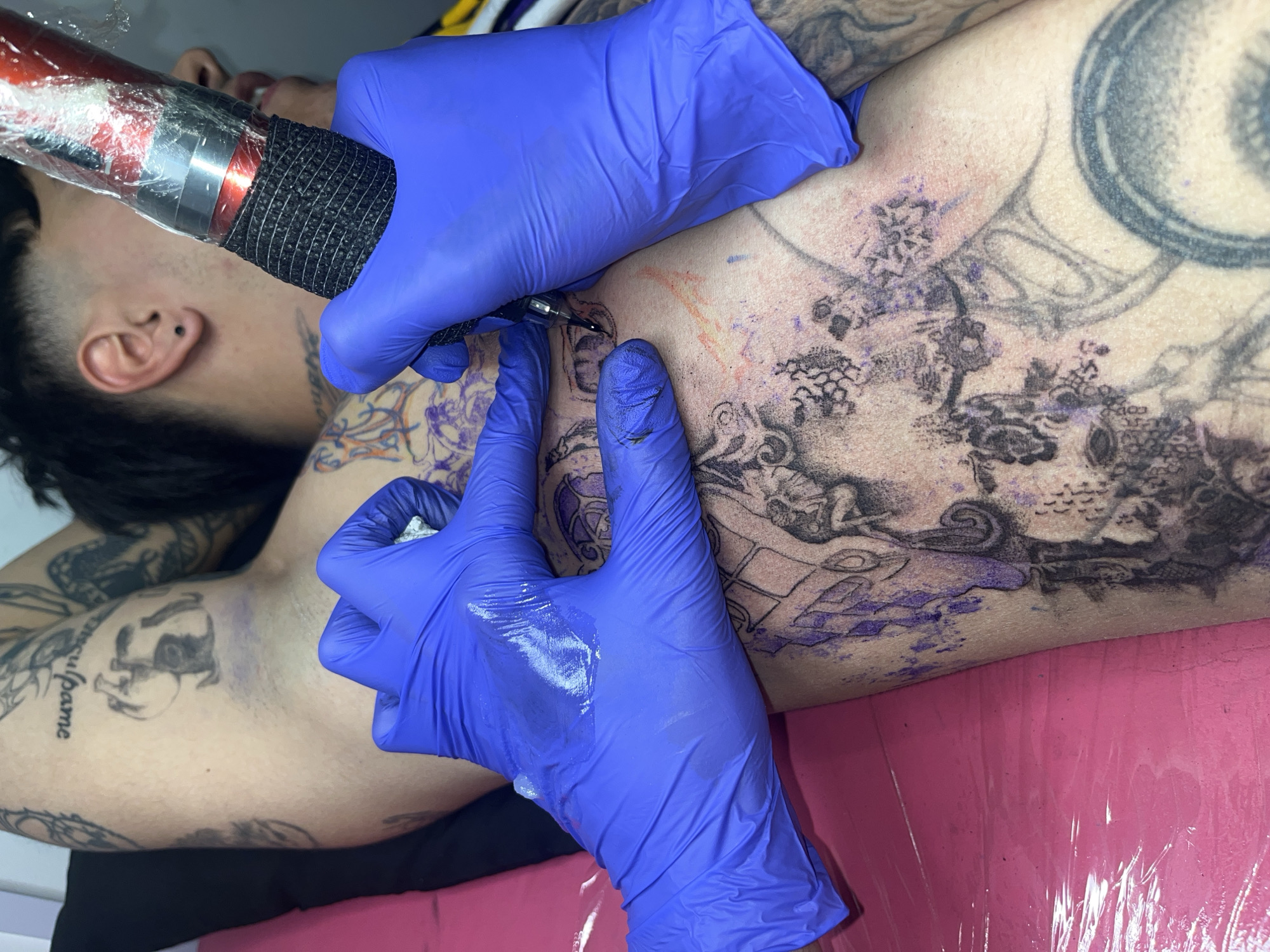 This close-up image captures a tattoo session in progress, likely within a tattoo studio in Buenos Aires, Argentina. A client, visible from the upper torso and right arm, lies on a pink padded surface, which is covered with clear plastic for hygiene. The client has dark hair and a small ear piercing.
The tattoo artist, represented by their hands covered in bright blue disposable gloves, is meticulously working on a new, intricate dark-ink design on the client's torso. They hold a red and black rotary tattoo machine, which is wrapped in clear protective plastic, actively applying ink to the skin. Distinct purple stencil lines are visible around the areas yet to be completed, indicating the ongoing outlining or detailing phase of the tattoo. The skin around the tattooed area shows some redness, a common reaction to the tattooing process.
The client's body is already adorned with several existing dark-ink tattoos. On their left bicep, partially visible text from an existing tattoo clearly reads "culpame," which is Spanish for "blame me." Other visible tattoos include a dark, abstract or figurative design on the lower left arm and intricate patterns across the upper torso. The client's partially visible face shows a neutral expression. The scene conveys a focused and professional tattooing environment.