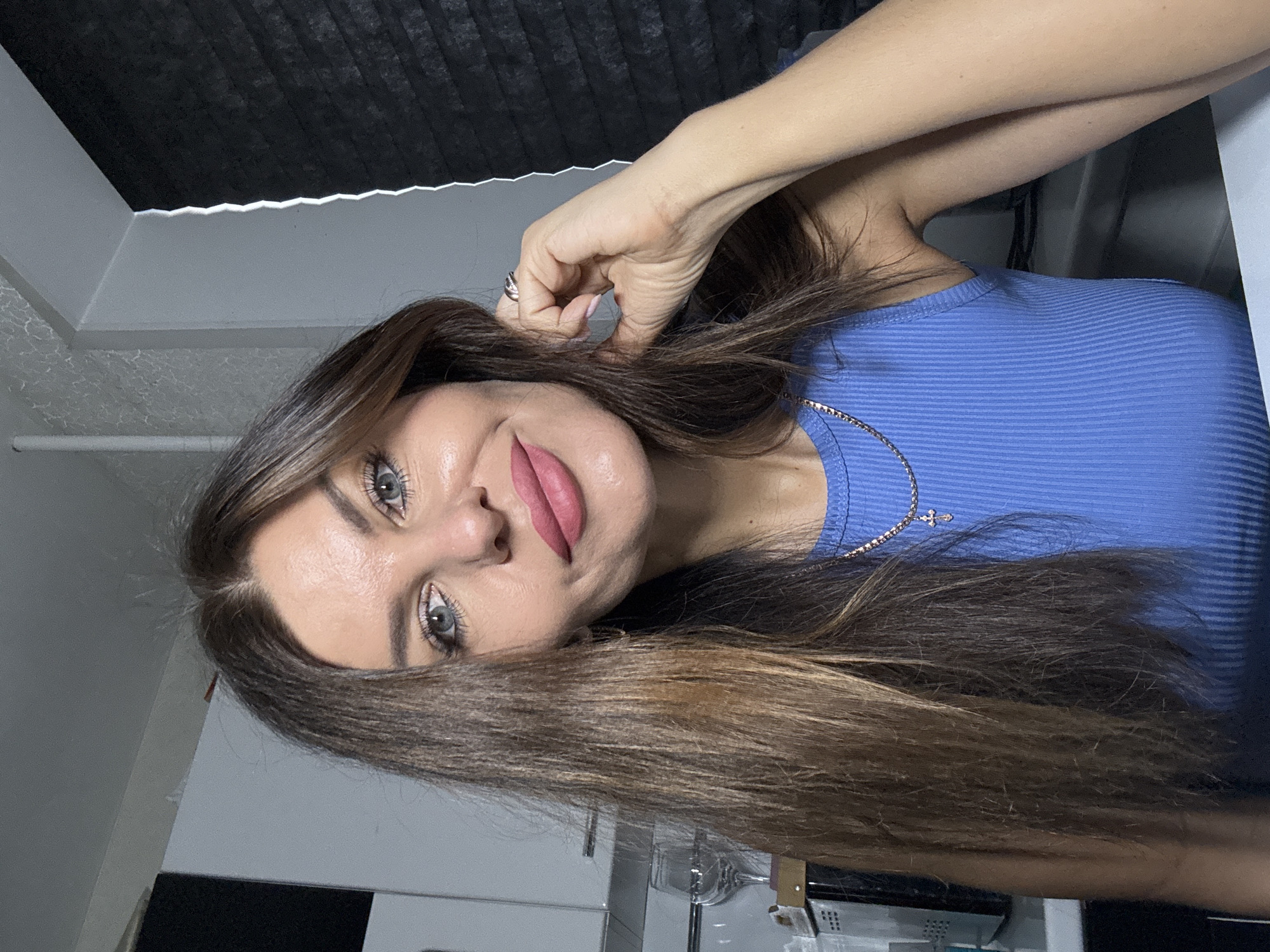 A woman is looking at the camera. She is wearing a blue top and a necklace. She has brown hair and light eyes. Her left hand is near her left ear. The background includes a window with dark blinds. The location is in an indoor setting, possibly in the city of Chaikovsky, Russia.