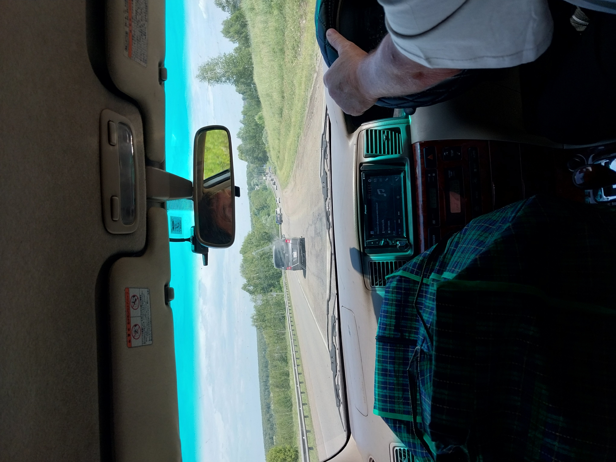 The image is taken from inside a car traveling on a road in Nalobikha, Russia. The perspective is from the driver's seat, looking forward and slightly to the right. The road is asphalt and appears to be in good condition, with a guardrail on the left side. There are other cars on the road in the distance. The landscape on either side of the road is rural, with green fields and trees under a cloudy sky.
Inside the car, the dashboard is visible, with a navigation system and various controls. A green and blue plaid blanket is draped over the passenger seat. The driver's hand is on the steering wheel. A side mirror reflects the driver's face and the passing scenery. The sun visors are up, and one has a warning sticker with Japanese text and symbols. The overall impression is of a road trip on a summer day.