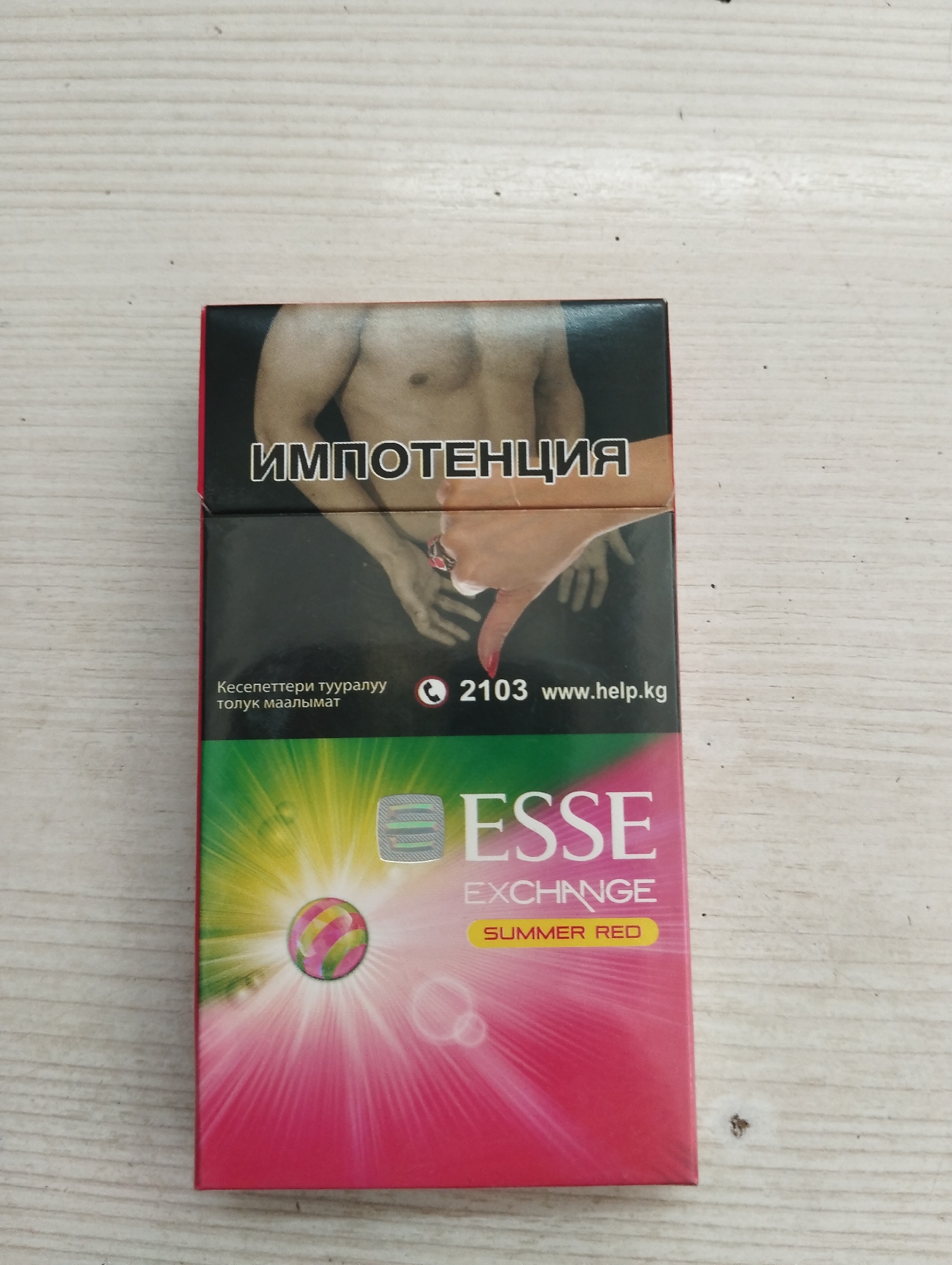 A close-up, slightly angled, top-down shot shows a pack of ESSE cigarettes placed on a light-colored wooden surface. The cigarette pack features a striking design with a gradient of pink and yellow, radiating outwards from a central circular graphic. The brand name "ESSE EXCHANGE" is prominently displayed in white lettering. Below this, "SUMMER RED" is written in yellow text on a yellow rectangular background. The upper portion of the pack displays the word "импотенция" (impotence) in large white Cyrillic letters on a black background, accompanied by an image of a man's bare torso. A hand with painted red nails is also visible reaching down towards the pack. The bottom left corner of the pack has text in Cyrillic, and to its right, a phone icon is followed by the numbers "2103" and the website "www.help.kg". A holographic sticker is visible in the upper right section of the colorful lower part of the pack. The wooden surface appears to be a tabletop or similar flat area, showing natural grain and some minor imperfections. The image is well-lit, with no shadows obscuring the details of the cigarette pack or its surroundings. The scene is static and appears to be an indoor photograph.