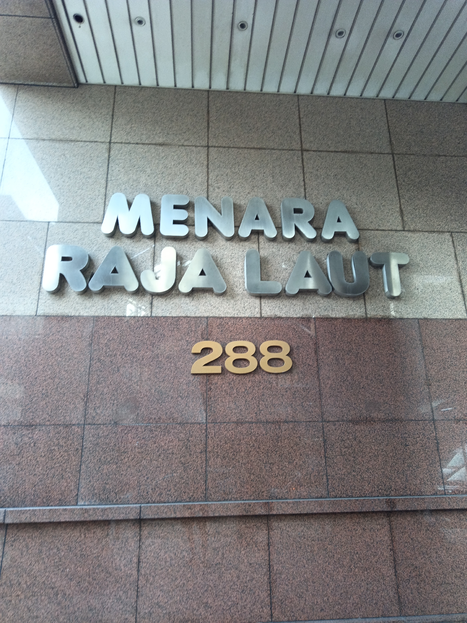 A close-up, eye-level shot features a building facade with metallic lettering that reads "MENARA RAJA LAUT" in large, silver, three-dimensional letters on the upper portion. Below this, in the lower portion, are the numbers "288" in gold, also three-dimensional. The background consists of textured beige tiles above the lettering and reddish-brown speckled granite tiles below. The image captures the building's name and address, suggesting it is a commercial or administrative building in Kuala Lumpur, Malaysia. The lighting indicates an outdoor setting during the day, possibly with some sunlight creating shadows and reflections on the metallic letters.