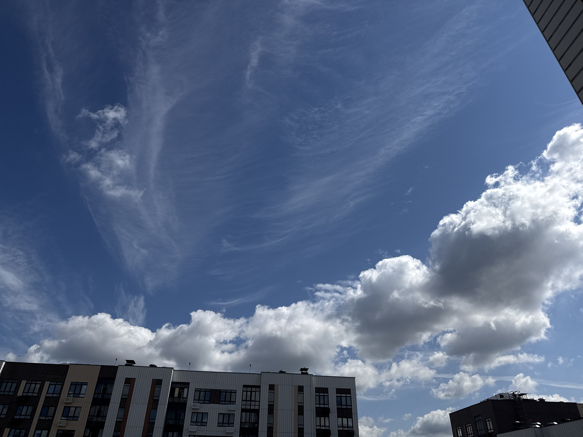 The image captures a bright, overcast sky above a residential area in Lyubertsy, Russia. The sky is a vibrant blue, filled with wispy cirrus clouds stretched across the upper portion of the frame, suggesting high-altitude atmospheric conditions. Lower in the sky, several puffy cumulus clouds are present, some appearing bright white in the sunlight, while others are beginning to show darker, gray undersides, indicating potential for precipitation.
Dominating the lower half of the image are the facades of modern apartment buildings. The main building in the foreground is a multi-story structure with a complex facade featuring a mix of dark, brown, and beige tiled sections. Rows of windows are visible, arranged in a grid pattern, with some windows appearing dark and others reflecting the bright sky. The building has a contemporary architectural style. To the right of this main building, a portion of another building is visible, darker in color and also featuring windows.
The perspective is looking upwards from ground level, showcasing the architecture against the backdrop of the sky. There are no people visible in the image, and no discernible actions are taking place. The environment depicted is urban, characterized by the presence of multi-story residential buildings under a dynamic sky. The overall impression is one of a typical day in a modern urban setting, with a focus on the interplay between architecture and the sky.