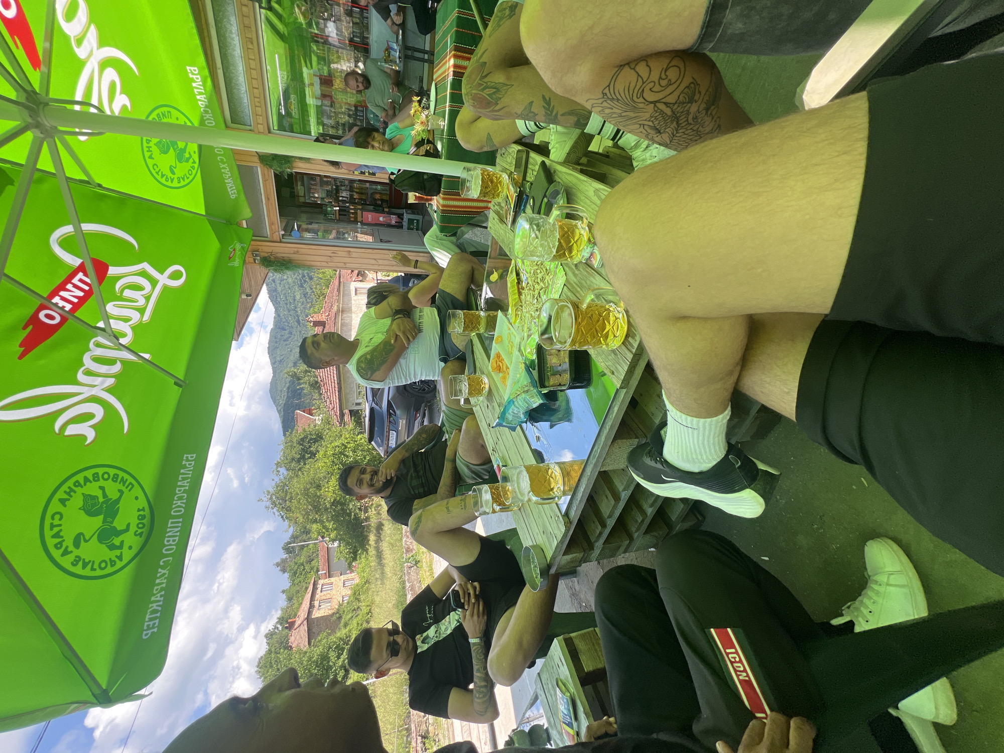 This image depicts an outdoor social gathering at a casual establishment in Касапите, Bulgaria, on a clear, partly cloudy day. The scene is framed by two large, green promotional umbrellas. One umbrella prominently displays the Cyrillic text "СТАРА ЗАГОРА" (Stara Zagora), "БЪЛГАРСКО ПИВО С ХАРАКТЕР" (Bulgarian Beer with Character), and a lion/cat logo with the year "1892". The other umbrella features "ЛИНФО" (LINFO) branding.
Approximately eight to ten adults, primarily males, are present. In the immediate foreground, the lower half of two individuals is visible: one male wears a black tracksuit with a red and green "ICON" stripe and white sneakers; another male, with tattooed legs, wears black shorts, white socks, and black Nike sneakers.
Around a rustic table constructed from wooden pallets, several men are seated. One man in a black t-shirt and sunglasses is focused on his smartphone. Another bearded man in a dark green t-shirt is smiling. A third man, with arm tattoos, wears a white t-shirt. Most individuals are dressed in shorts and short-sleeved shirts, indicating warm weather. The table holds multiple glass mugs filled with a light-colored liquid (consistent with beer), bowls containing snacks such as chips and pretzels, and personal items like smartphones and an ashtray.
In the mid-ground, a storefront, likely a small bar or café, with glass doors and windows, reveals shelves stocked with bottles inside. An older woman in a green top and two additional men are visible within the establishment. Beyond, the background features rural houses with red-tiled roofs, dense green foliage, and distant hills or mountains under a partly cloudy blue sky. A dark-colored SUV is partially visible parked behind the group. The overall ambiance is relaxed and social.