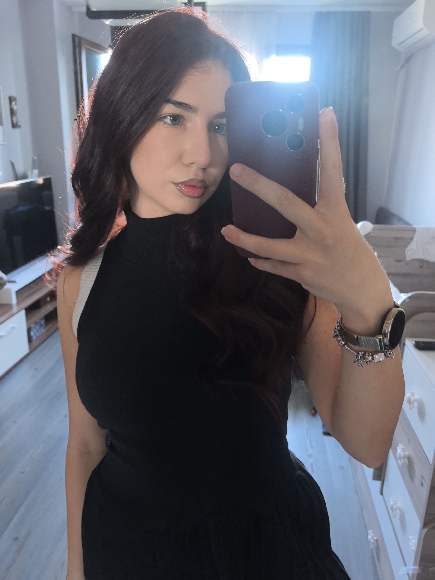 A young woman is taking a selfie in what appears to be an apartment. She is holding a smartphone in her right hand, obscuring part of her face. She is wearing a black turtleneck dress or top. The background includes a television, furniture, and a window with sunlight streaming through. She has a watch and bracelet on her right wrist. The location is Unknown, Unknown.