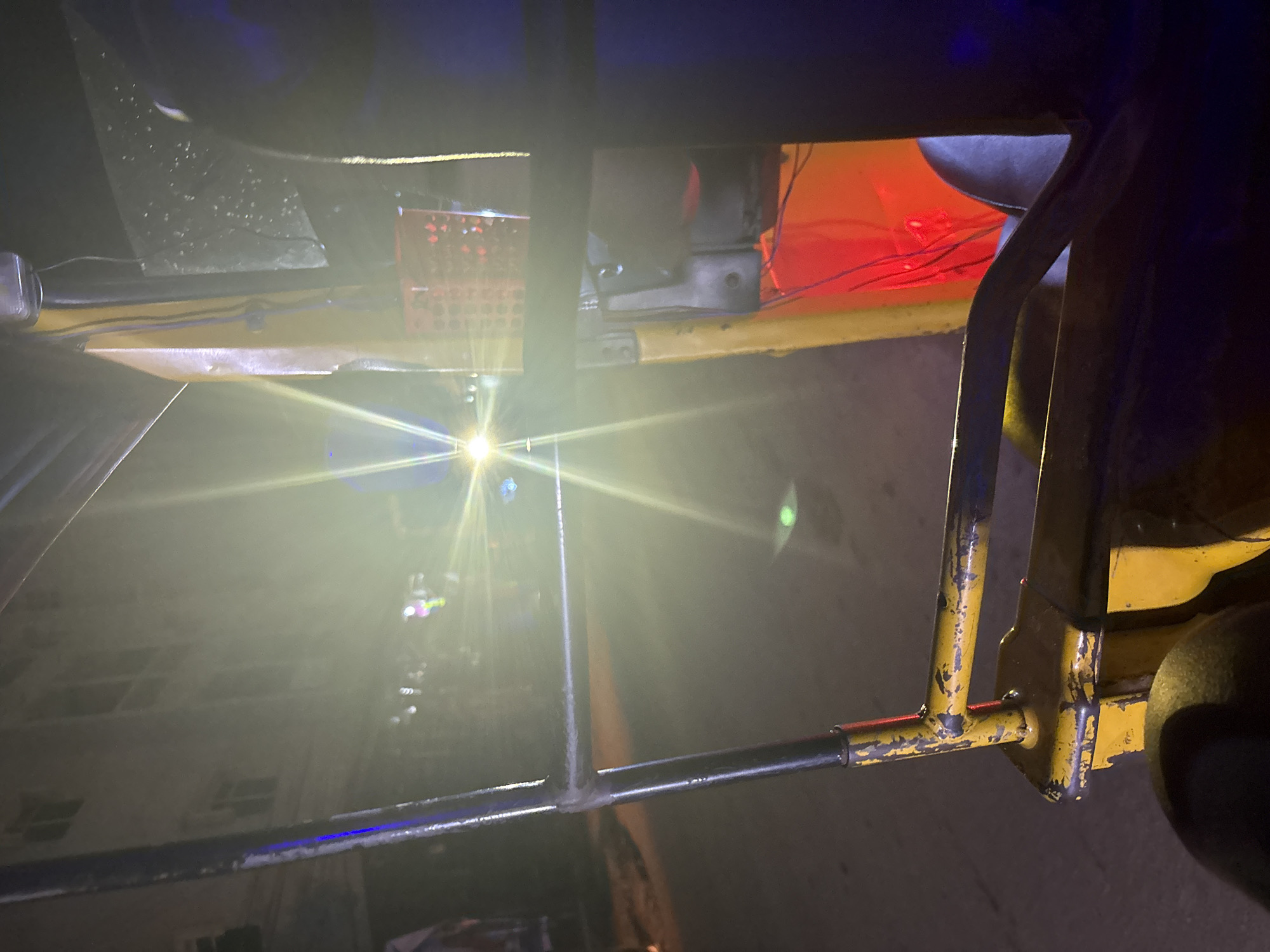 The image shows the interior of a yellow auto-rickshaw at night, likely in Nkpor, Nigeria. Bright headlights from another vehicle create a strong glare in the center of the image. The auto-rickshaw has a yellow frame with black metal bars. Visible inside are a red object, possibly a basket or container, and some electrical wiring. A person's leg and dark clothing are also visible on the right side. The background is dark with some indistinct lights and the facade of a building. The scene appears to be on a paved road.