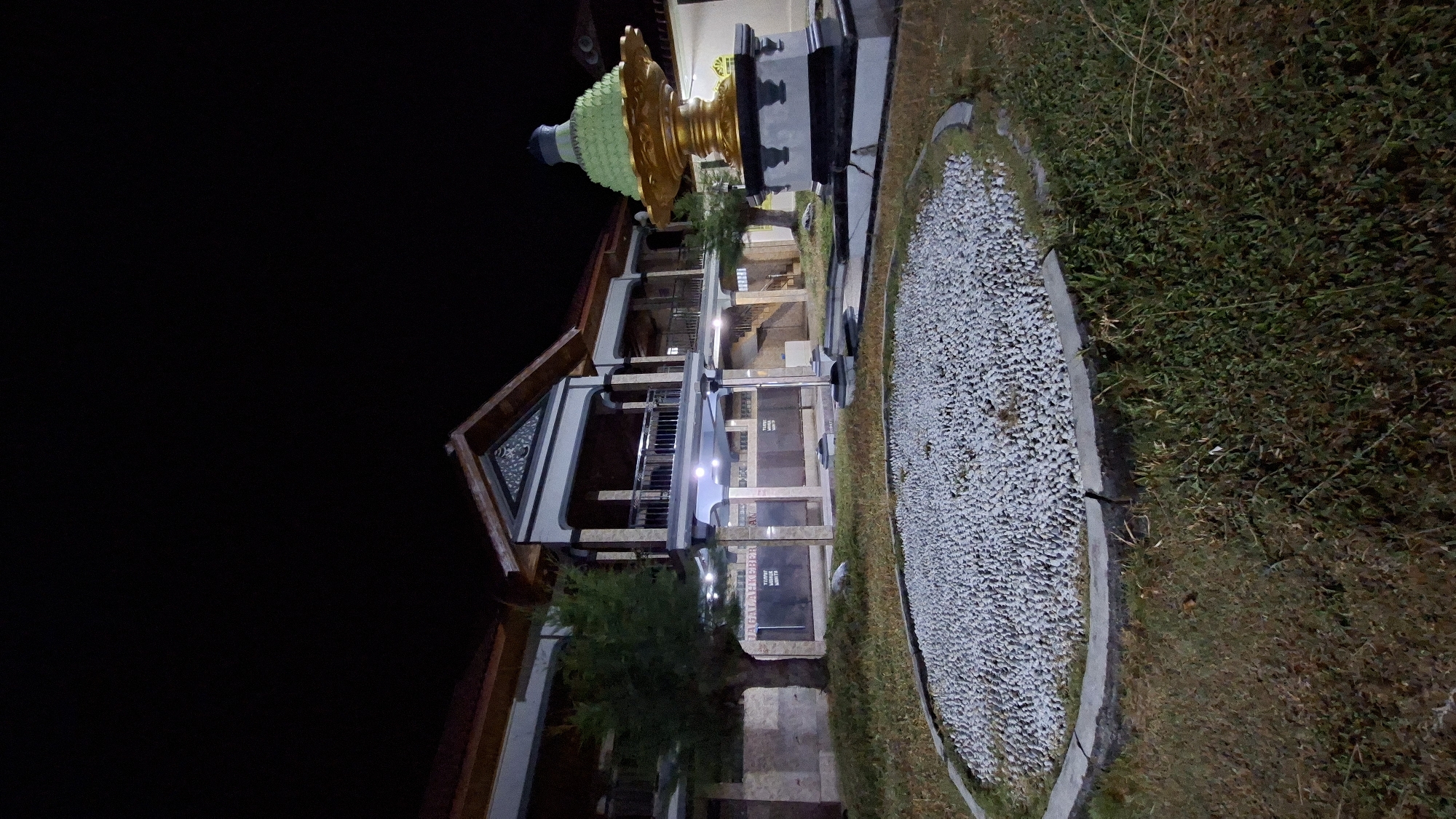 This is a medium-shot, eye-level, vertical, nighttime photograph of a building and its exterior surroundings in Lhokdagang, Indonesia. The building appears to be two stories with a dark roof and light-colored walls. It has a covered balcony area on the second floor with a railing. Decorative architectural elements are visible on the facade. In the foreground, there is a circular garden bed filled with small, light-colored stones, bordered by a concrete edge. To the right of the stone garden, there is a dark, ornate fountain or monument with a green, layered top. The ground is covered in sparse grass and some dry vegetation. The lighting in the scene appears to be artificial, possibly from streetlights or the building itself, creating shadows and highlighting certain features. No people are visible in the image. The overall impression is one of a quiet, well-maintained public space at night.