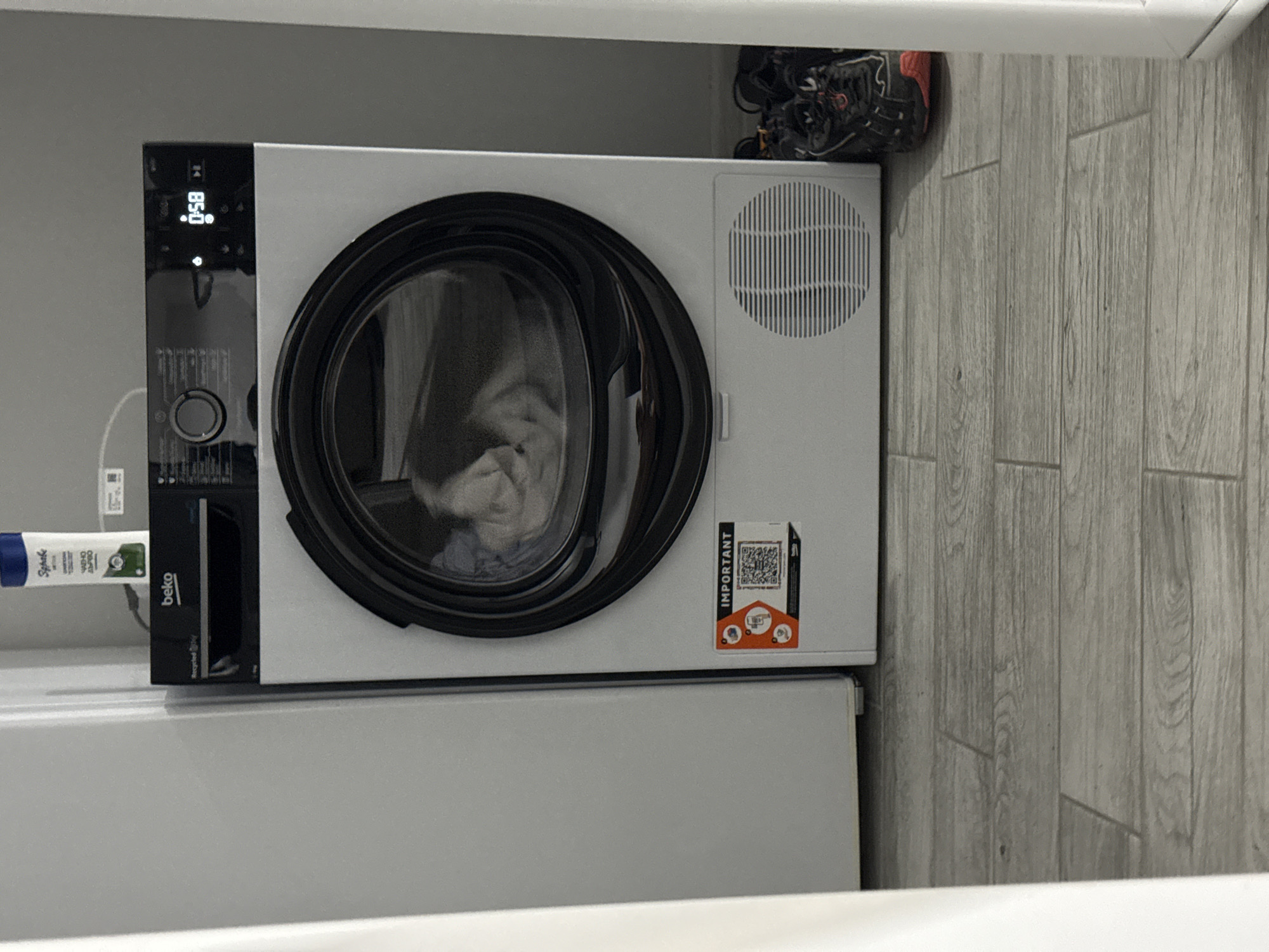 A Beko dryer is centrally located in a laundry room, its door slightly ajar, revealing laundry inside. A white bottle of what appears to be shampoo or conditioner stands on top of the dryer. A gray wooden plank floor is visible beneath the dryer. To the right of the dryer, a pair of black and orange athletic shoes are placed. The room appears to be part of a residential setting, likely a home or apartment. No specific location in Stara Zagora, Bulgaria is identifiable from the image.