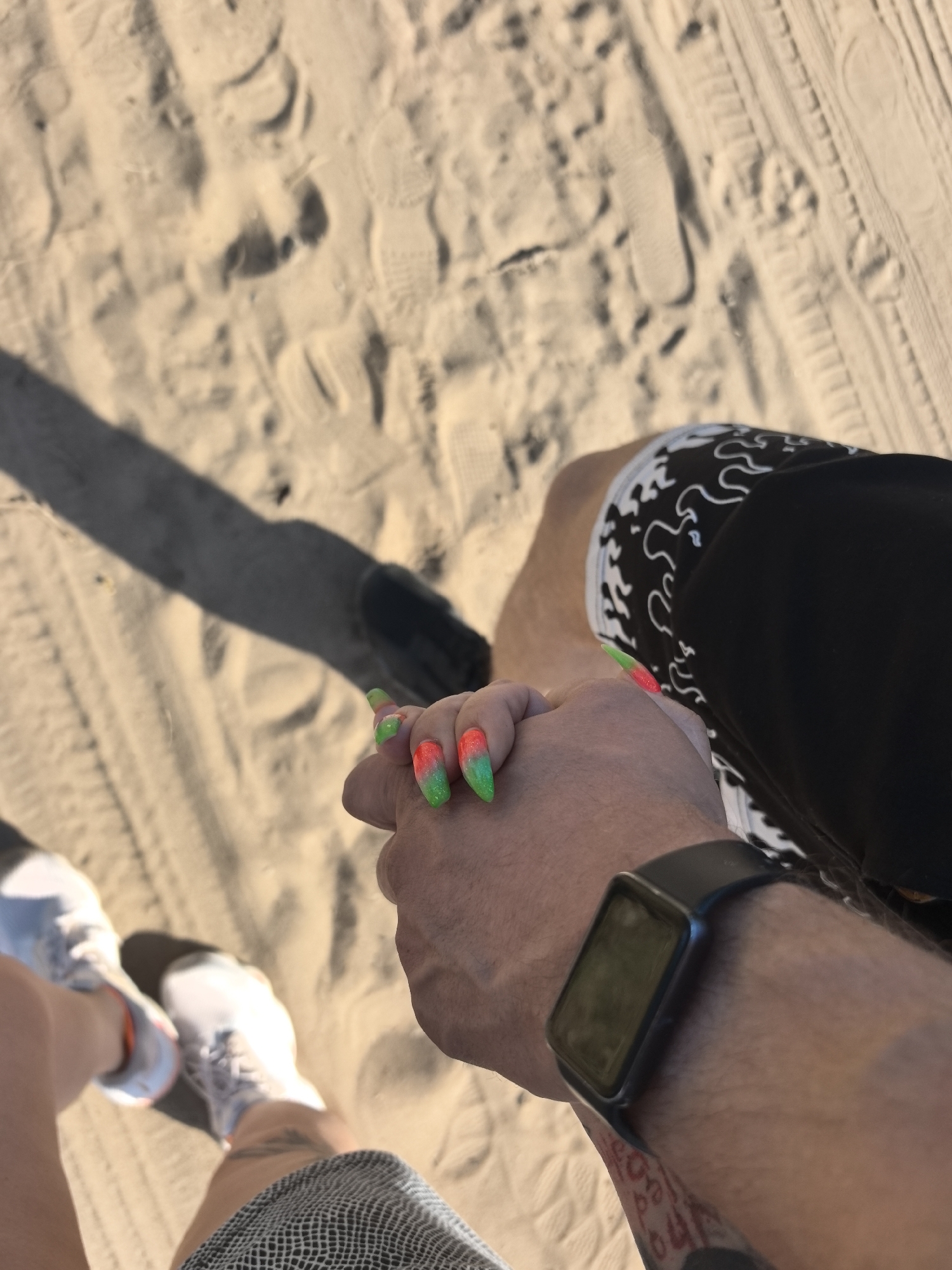 A couple is holding hands on a beach. The sand has many footprints and tire tracks. The person on the right is wearing black shorts with a white pattern, and a black watch. The person on the left is wearing white sneakers and gray shorts. The person on the right has a tattoo on their arm, and the person on the left has nails painted red and green. The photo was most likely taken on a beach somewhere in the world, not specifically in Noviy, Russia, as there is no identifying information to place it there.