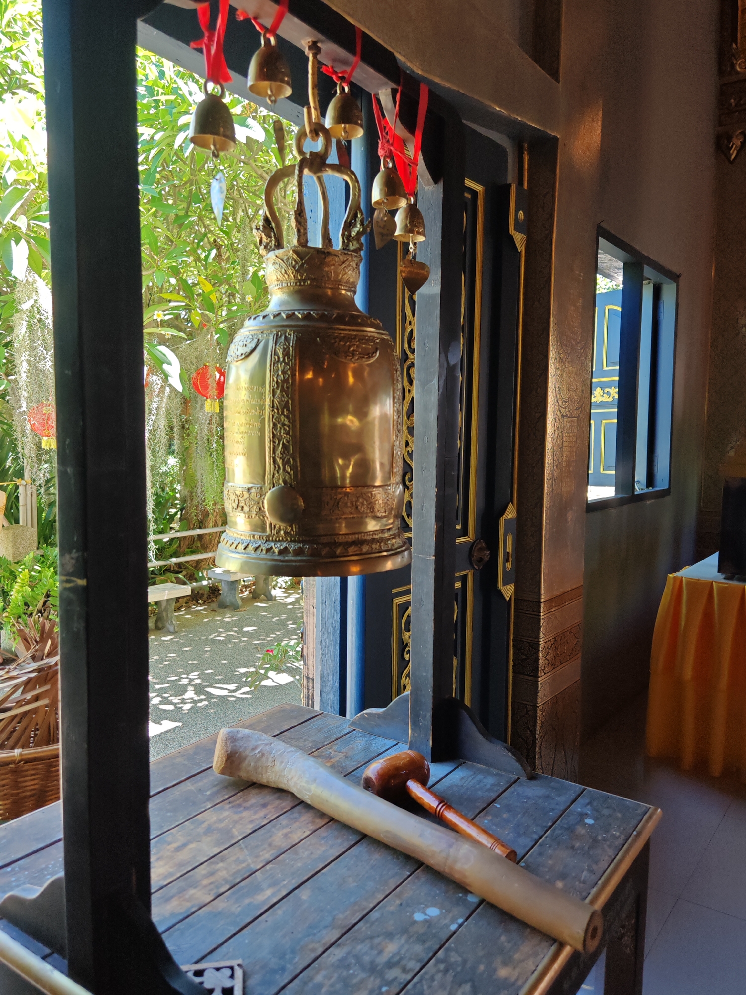 The image depicts a large brass bell hanging inside a structure, likely a temple or religious site, in Ban Bang Rak, Thailand. The bell is ornate with inscriptions and is suspended from a wooden beam by a metal bracket. Several smaller brass bells are also visible, hanging from red ribbons above the main bell. In the foreground, a wooden table holds two striking implements: a long, cylindrical object, possibly made of bamboo or wood, and a smaller wooden mallet with a red handle. Behind the bell, an open doorway reveals a sunlit garden area with lush greenery, hanging decorative lanterns, and stone benches. The interior of the structure features dark blue doors with gold trim, and a window with blue panes. A yellow tablecloth covers a table in the background. The scene is well-lit, suggesting daytime.