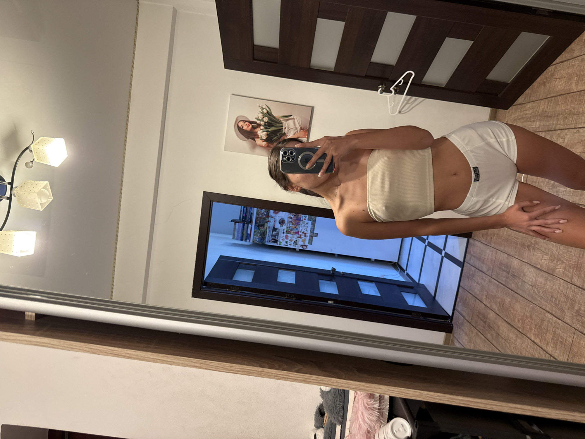 A young woman with dark hair, wearing a beige crop top and white shorts, stands in a room and takes a selfie in a large mirror. The room has wooden flooring and dark wood doors. In the reflection, a door is ajar, revealing a hallway with a tiled floor. On the wall, a framed picture of a woman holding a bouquet of flowers is visible, as is a light fixture on the ceiling. The overall impression is that of a private dwelling.