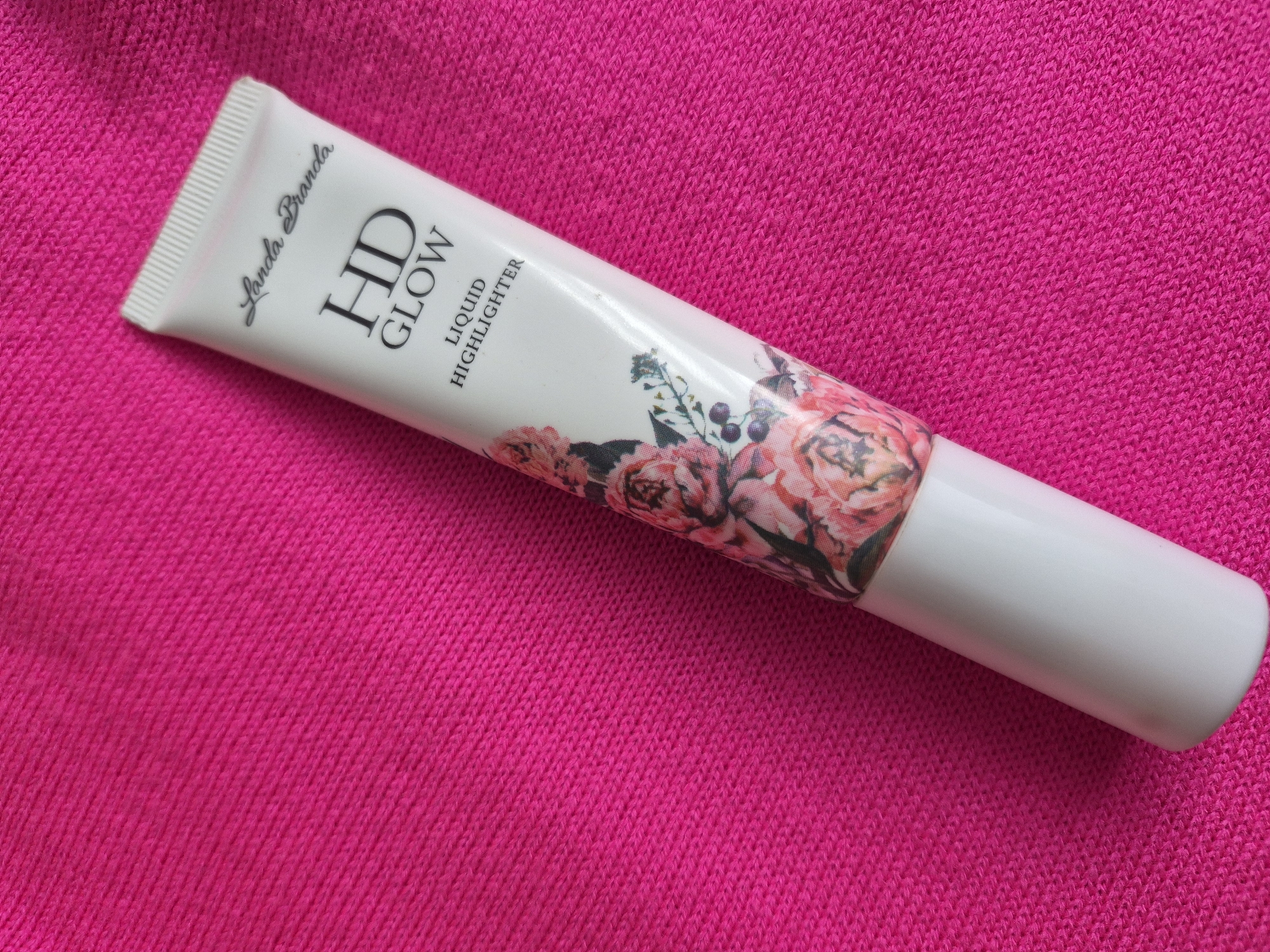 A tube of "Landa Branda HD Glow Liquid Highlighter" is on a hot pink knitted background. The tube is white with black text. The tube has a floral design.