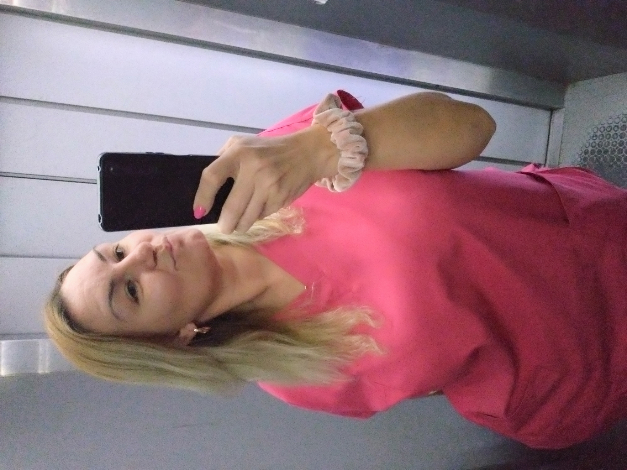 A woman is taking a selfie in what appears to be an elevator. She is wearing a pink top and a scrunchie on her wrist. Her fingernails are painted pink. The background consists of the elevator walls, which are metallic. It is not possible to ascertain the location in the city of Stara Zagora, Bulgaria, from this image.