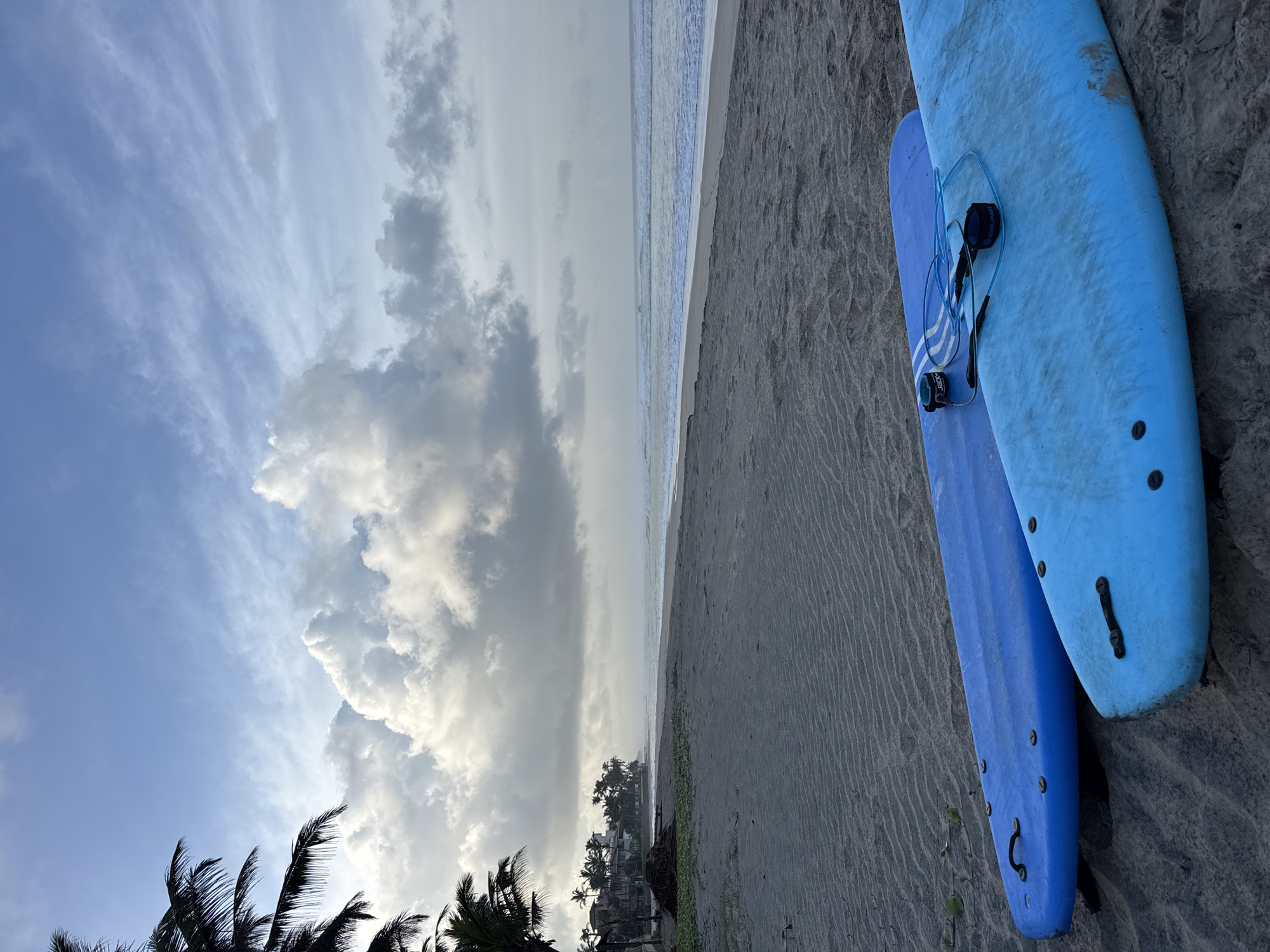 This image depicts a tranquil beach scene in Kathaluwa, Sri Lanka, under a partially cloudy sky. In the foreground, two bright blue surfboards are resting on the sandy beach. The larger, lighter blue surfboard is closer to the viewer and occupies the bottom right portion of the frame. The second surfboard, a slightly darker shade of blue, is positioned behind and to the left of the first. Both surfboards appear to be in good condition and are oriented parallel to the waterline.
The beach itself is composed of fine, gray sand, with subtle ripples indicating the gentle ebb and flow of the tide. The ocean stretches out to the horizon, its surface reflecting the muted light of the sky. Gentle waves are breaking along the shore, creating a thin line of white foam.
In the middle ground, a line of palm trees and some buildings can be seen along the coastline, suggesting a developed area adjacent to the beach. The vegetation is lush and tropical, characteristic of the Sri Lankan coast. The sky is a mix of soft blue and dramatic, billowing white clouds, particularly prominent in the upper half of the image. The overall atmosphere is serene and peaceful, evoking a sense of a quiet morning or late afternoon at a tropical destination. There are no people visible in the scene.