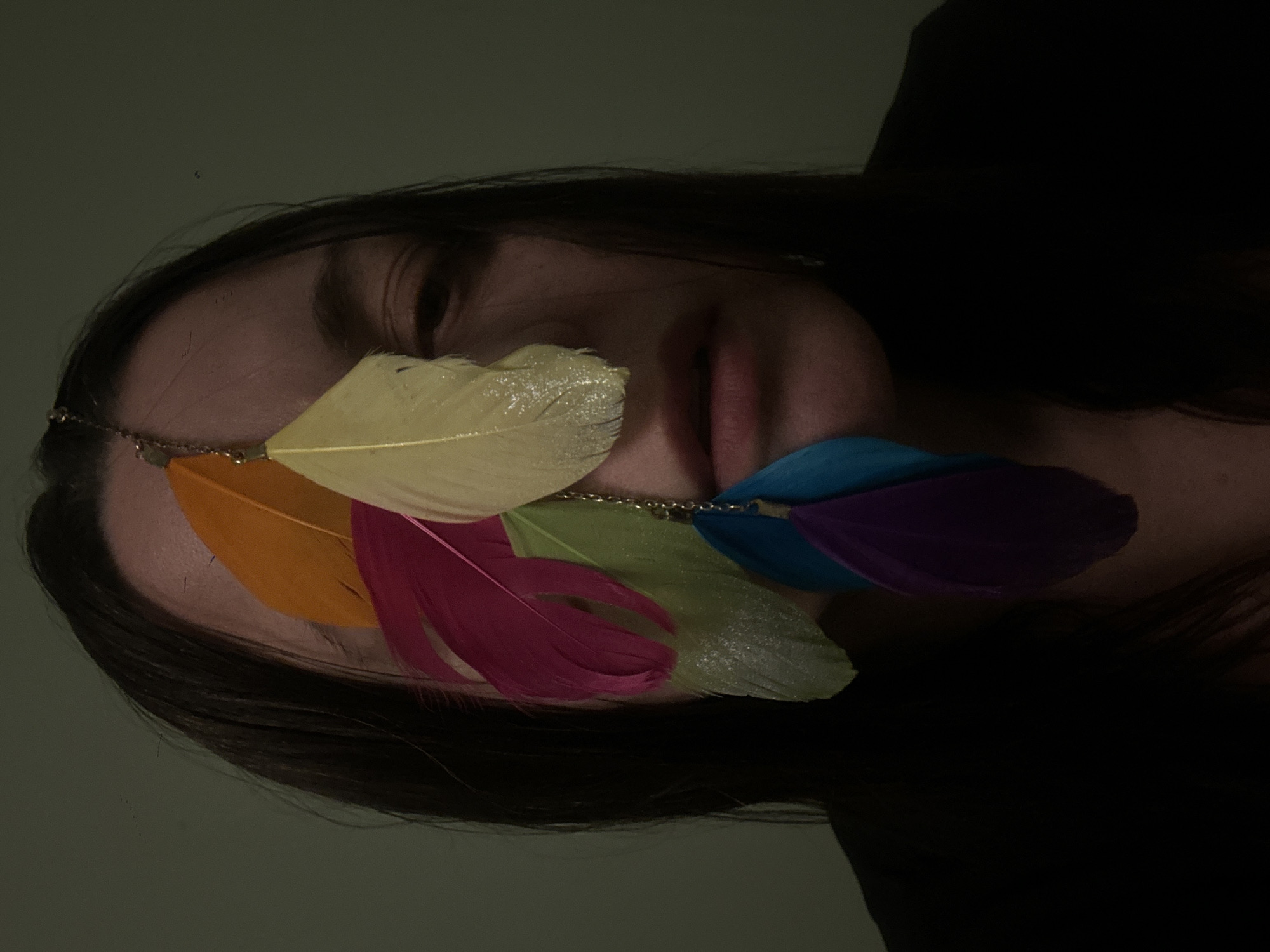 A close-up, dimly lit portrait features a person's head and face, turned slightly to their left. The eyes are obscured by a decorative chain adorned with multiple vibrant feathers. The feathers are colored yellow, orange, hot pink, light green, blue, and purple, arranged across the upper part of the face, covering the eye area. The person has long, dark hair visible along the side of their face and draped around the head. Their lips are slightly parted. The background is a plain, dark greenish-grey surface, suggesting an indoor setting. The low light creates a soft, somewhat mysterious ambiance. There is no specific activity depicted beyond posing for the portrait. No text is visible in the image.