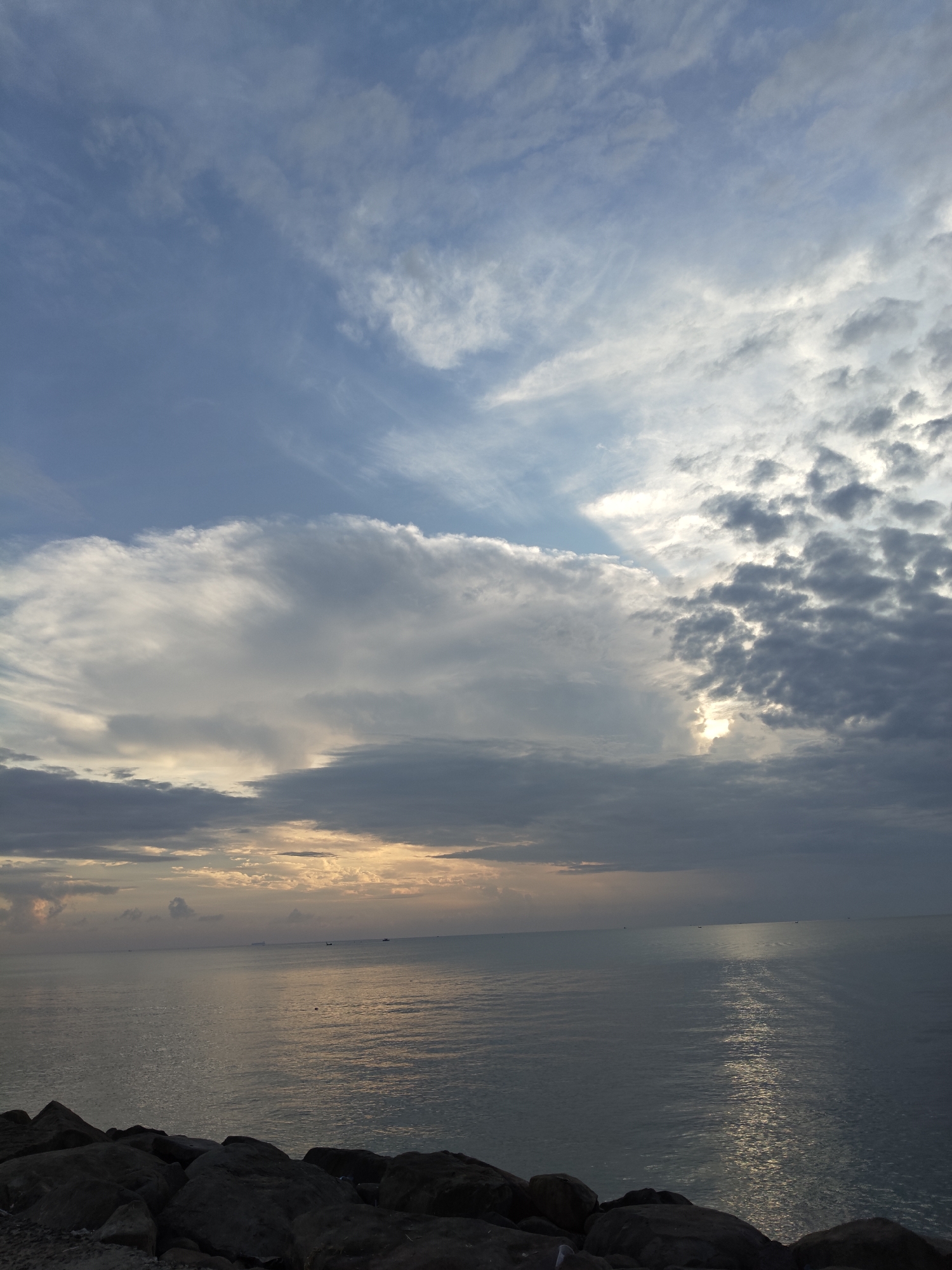 This is a wide-angle, eye-level, photograph of the ocean at Lhokseumawe, Indonesia. The sky is filled with a variety of clouds, ranging from wispy cirrus formations to larger, more ominous cumulonimbus clouds. The sun is partially obscured by the clouds, casting a warm glow across the water. The ocean itself is calm, with gentle ripples reflecting the light from the sky. In the foreground, large rocks line the shore, suggesting a rocky beach or a breakwater. The overall impression is one of a serene and beautiful seascape, possibly at sunrise or sunset. There are no people or animals visible in the image. The horizon is visible, with a few distant ships or boats barely visible on the water. The location is confirmed to be Lhokseumawe, Indonesia.