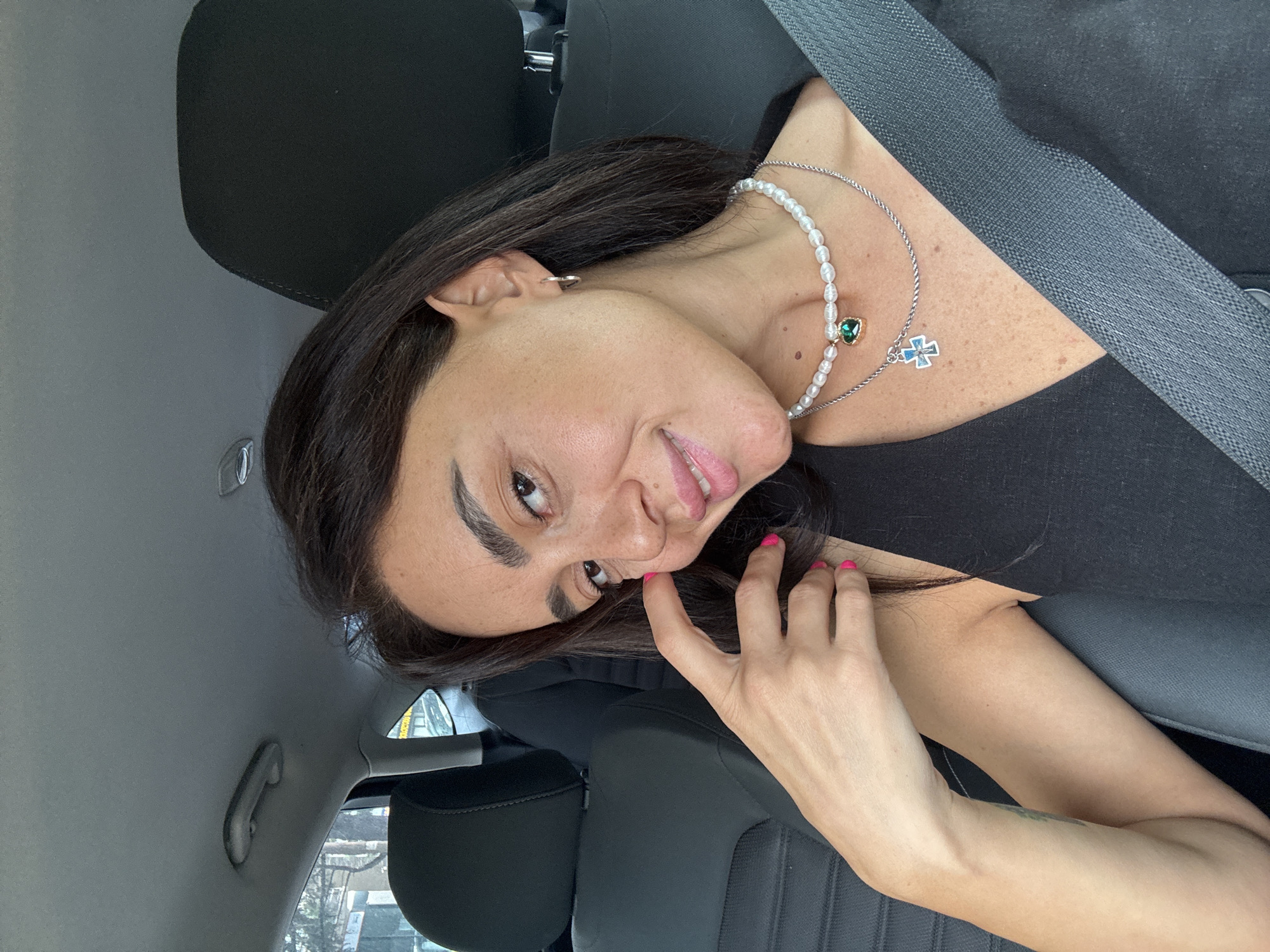 The image is a selfie taken from inside a car, with the subject seated in the driver's seat. The woman has dark hair, brown eyes, and is wearing a black top, a pearl necklace with a green heart pendant, and a silver chain with a cross pendant. Her fingernails are painted bright pink. She is touching her cheek with her right hand and smiling. The interior of the car is visible, including the headrest of the passenger seat, a seatbelt, and parts of the dashboard and ceiling. The background outside the car window shows a blurred view of trees and buildings, suggesting an urban environment. The overall lighting suggests it is daytime.