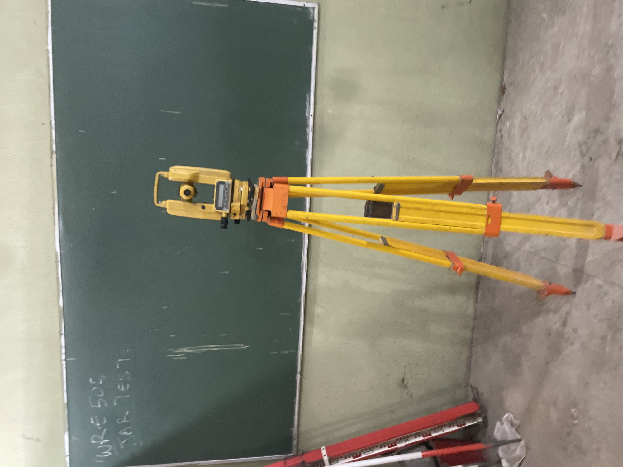In a classroom or laboratory environment located in Ihiagwa, Nigeria, a surveying instrument, specifically a total station mounted on a yellow tripod with orange accents, is positioned in the foreground. The instrument is facing towards the viewer, its digital display visible. Behind the tripod, a dark green chalkboard is mounted on a light green wall. White chalk writing on the chalkboard is partially visible, reading "WRR 505" and "JAR TEST.". To the left of the tripod, a red and white surveying staff leans against the wall, partially obscured. The floor is a concrete surface, appearing somewhat worn. The lighting in the scene is moderate.