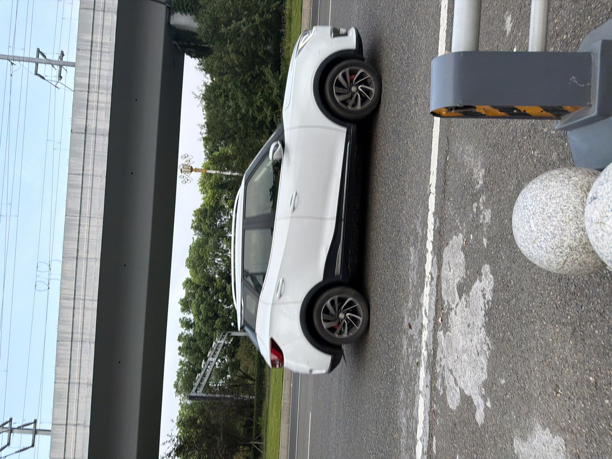 The image shows a white SUV driving on a paved road in Jiangjin, China. The car is in motion, as indicated by the slight blur. Overhead, a concrete overpass is visible, with power lines and poles in the background. Lush green trees line the side of the road. In the foreground, the road surface shows some wet patches and a white line. A gray traffic barrier with yellow and black reflective tape is positioned on the right side of the image, along with two large, spherical granite bollards. The sky is overcast, suggesting a cloudy day.