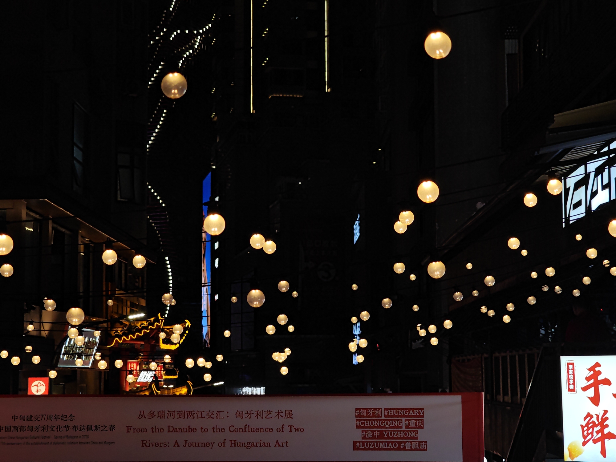 The image shows a street scene in the Yuzhong District of Chongqing, China, illuminated by numerous hanging spherical lights. A banner in the foreground advertises a cultural exhibition titled "From the Danube to the Confluence of Two Rivers: A Journey of Hungarian Art," commemorating the 77th anniversary of diplomatic relations between China and Hungary. The banner includes the hashtags #HUNGARY, #CHONGQING, #YUZHONG, and #LUZUMIAO. The scene includes a blend of modern buildings and traditional Chinese architectural elements. A sign with Chinese characters for "Fresh Dumplings" is visible on the right. The photo captures an evening atmosphere.