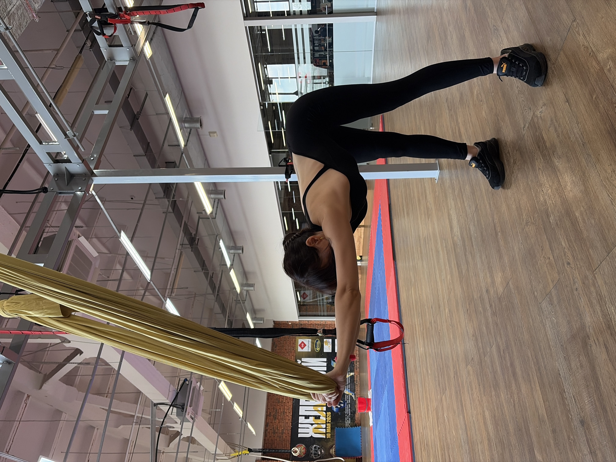 In a gymnasium in Chaikovsky, Russia, a woman in black athletic wear and black Puma sneakers is performing an aerial yoga stretch. She is holding onto a yellow aerial silk hammock, her body is bent at the waist with her arms extended forward, and her head is lowered towards the floor. The gym features a wooden floor, red and blue mats on the ground, and industrial-style lighting and metal structures on the ceiling. Posters with Russian text are visible on a brick wall in the background.