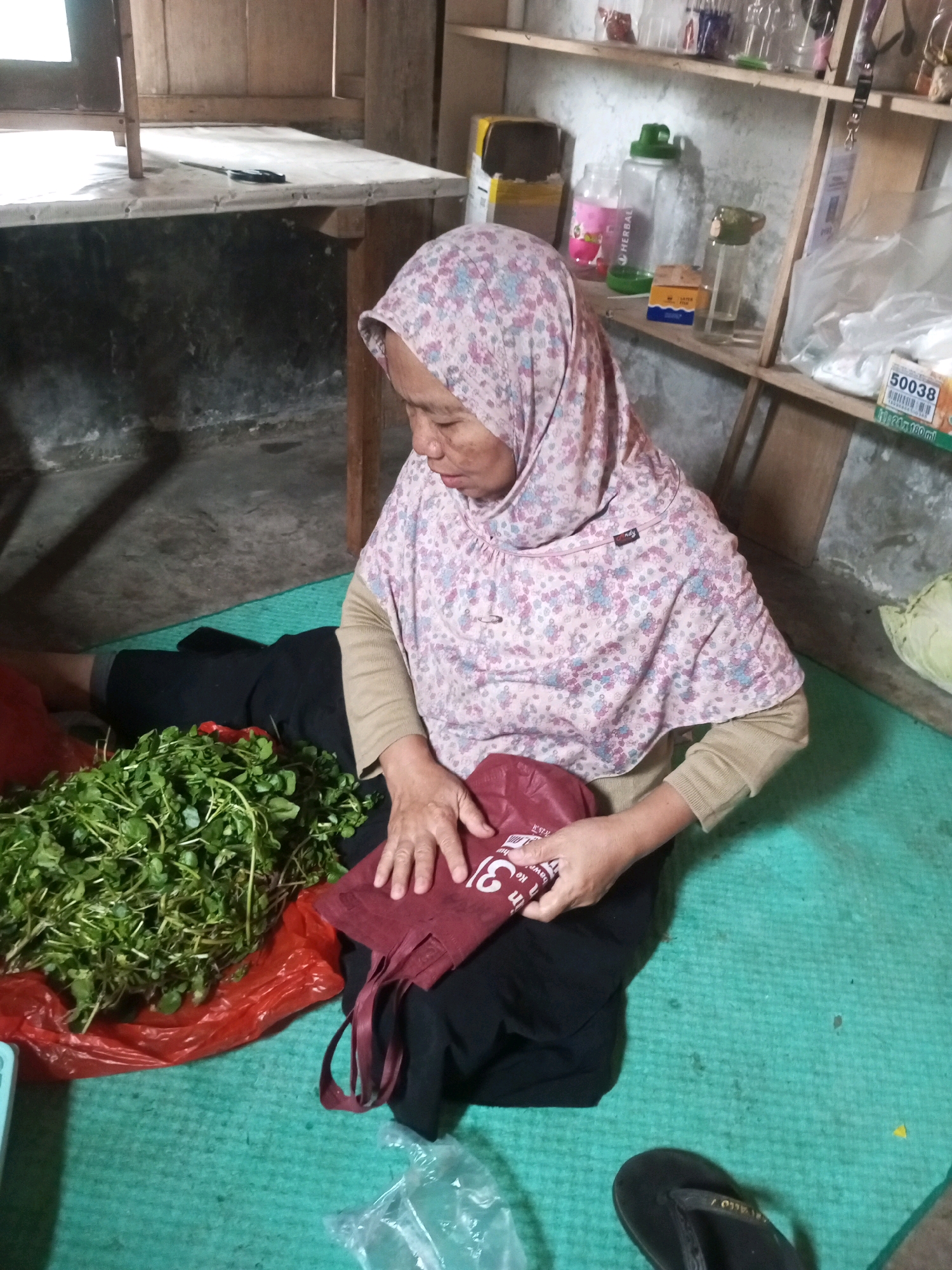In Kalibeber, Indonesia, a woman wearing a pink hijab and a long-sleeved tan shirt is sitting on a green mat. She is hunched over a large bunch of green leafy vegetables, which are placed on a red plastic bag. Her hands are manipulating a dark red cloth bag with white lettering. Behind her, shelves are filled with various bottles and containers. A wooden table with a white cloth covering is visible in the background, with a knife resting on it. The environment appears to be indoors, possibly a home or a market stall, with a rustic wooden structure forming the walls and ceiling.