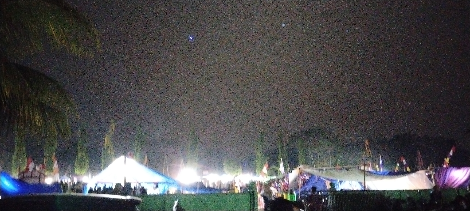 This is a nighttime photograph taken in Sido Rahayu, Indonesia, depicting an outdoor event. The sky is dark with visible noise, and there are a few faint celestial bodies or distant lights. In the foreground, the fronds of a palm tree are visible on the left side. The middle ground is occupied by a gathering with multiple tents and stalls, illuminated by artificial lights, creating a festive atmosphere. Some of these structures appear to be white or blue tents. Several flags, some red and white, are planted around the event area. In the background, a dense line of trees forms a dark silhouette against the sky. The ground is not clearly visible but appears to be covered in some sort of green material or grass in the lower portion of the image. The overall impression is of a community event or festival taking place after dark.