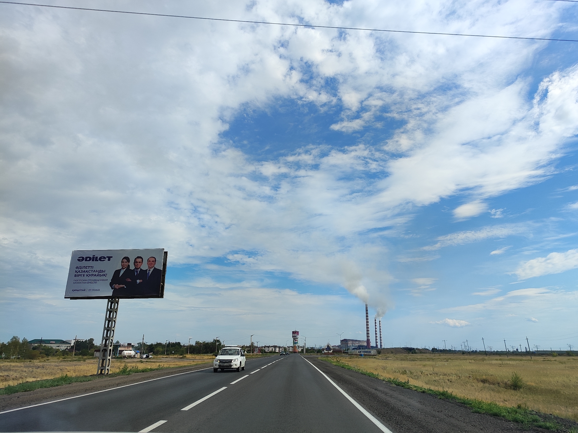 The image captures a view from a vehicle traveling on a highway in Aksu, Kazakhstan. On the left side of the road, a large billboard displays three individuals in formal attire and text in Kazakh. The environment is characterized by a vast, open landscape with dry, yellowish grass and scattered trees. In the distance, industrial structures, including tall smokestacks emitting white smoke, are visible. Power lines crisscross the sky, and a few buildings and vehicles are present along the roadside. The sky is partly cloudy, with a mix of white clouds against a blue backdrop. The road itself is paved with white lane markings, and a white car is driving in the same direction as the viewer.