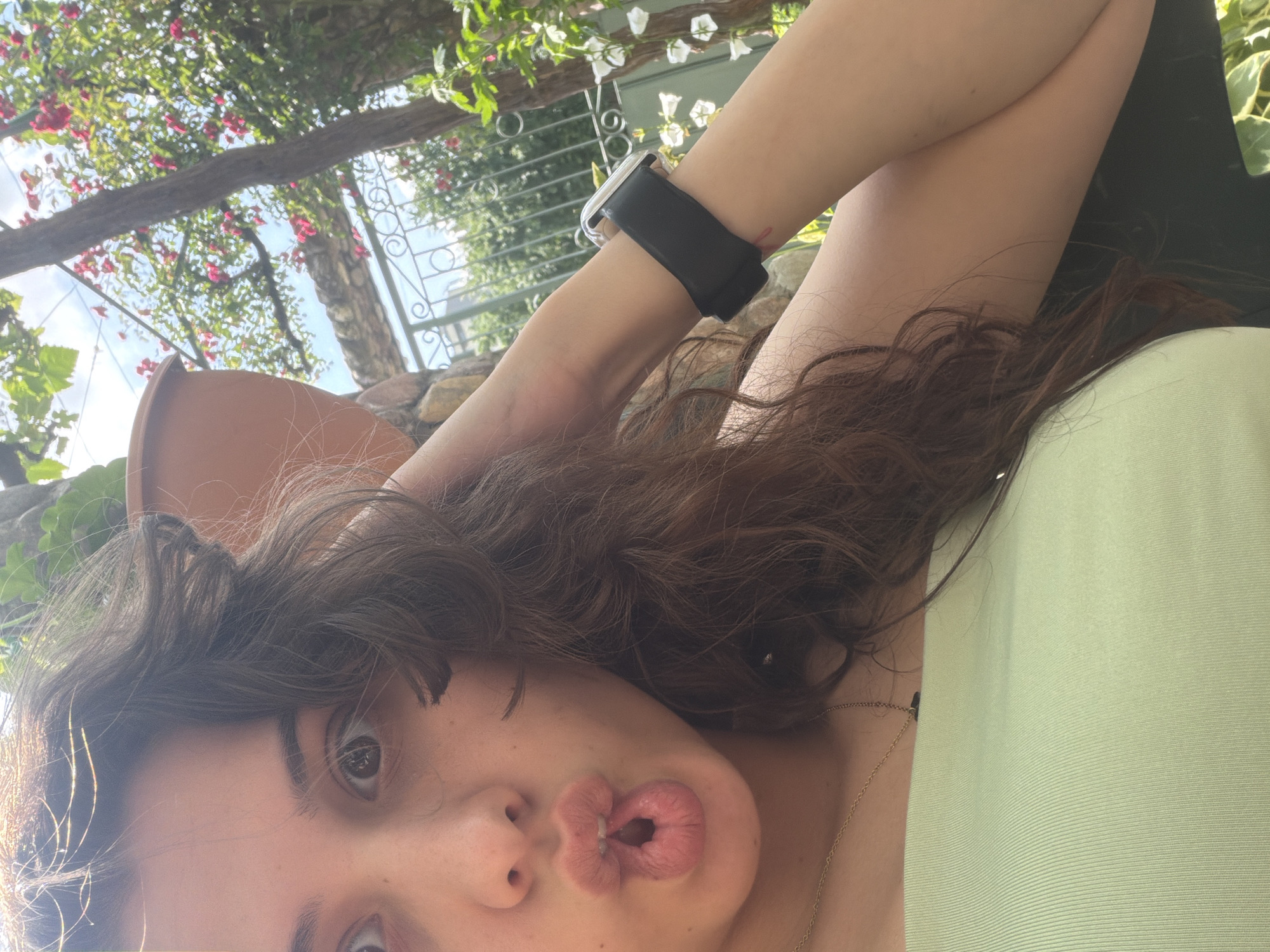A selfie-style photograph captures a person with a playful expression, appearing to lie down or recline outdoors. The main subject is a young person with dark, wavy hair, looking upwards towards the camera with wide eyes and pursed lips. They are wearing a light green top and a thin gold chain necklace. A smartwatch with a black band is visible on their right wrist, which is bent with the arm raised.
The background suggests an outdoor setting, possibly a garden, patio, or pergola. Above the person, there's an overhead structure covered in green foliage and flowering vines, featuring both red and white blossoms, likely in bloom under a bright, sunny sky with some clouds. A large terracotta pot is partially visible to the left of the person's head. Further in the background, a stone wall or structure is present, along with a decorative dark metal railing or trellis. The lighting indicates it is daytime, likely mid-morning or afternoon, with bright sunlight filtering through the leaves. No visible text is present in the image.