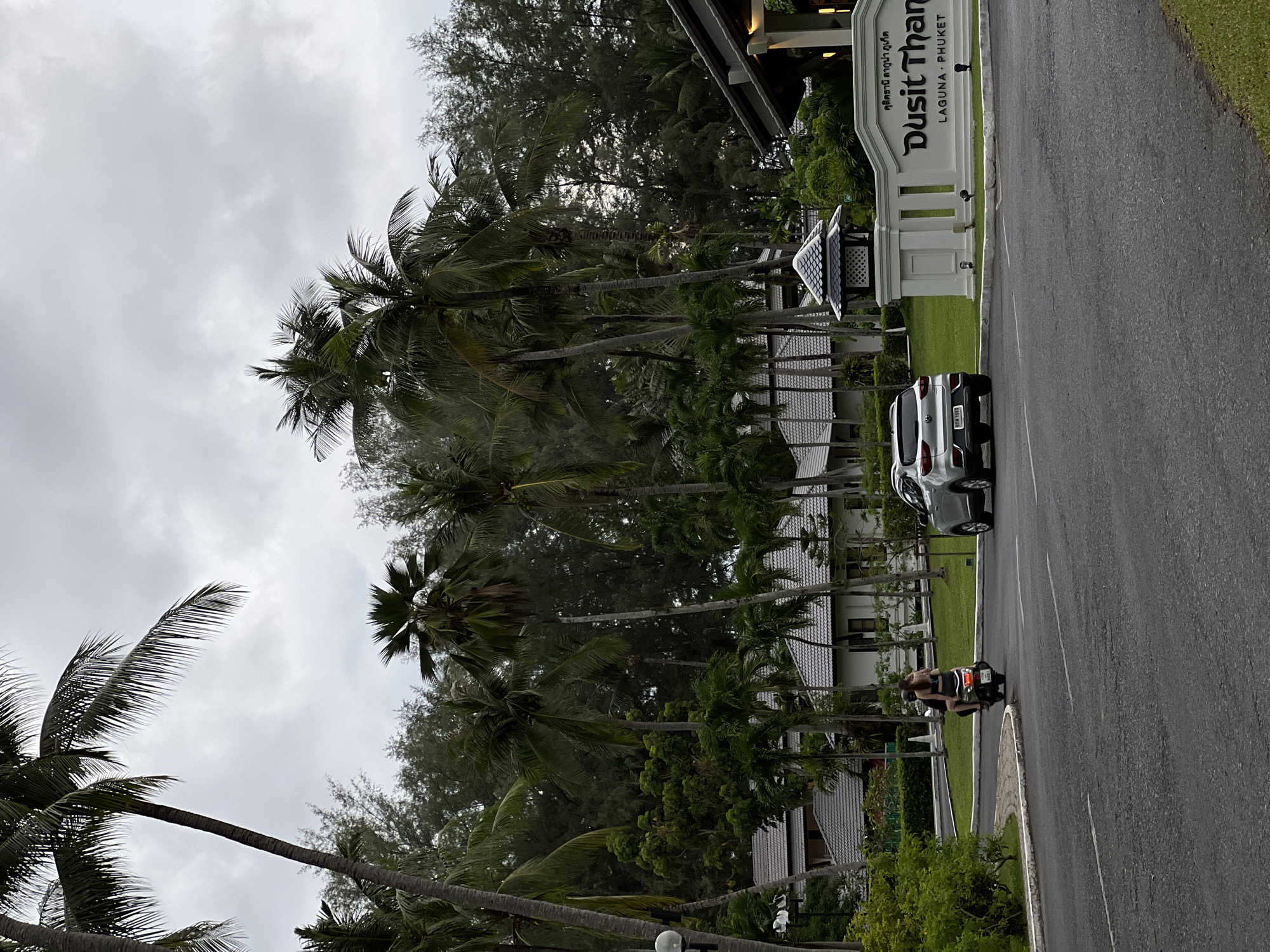 A silver SUV is parked on the right side of a paved road. A person is riding a scooter with a passenger in the center of the road, heading away from the camera. Both individuals on the scooter are wearing casual clothing. To the left of the road, there are several buildings with white walls and grey roofs, surrounded by lush green trees and palm trees. A white sign with dark lettering that reads "Dusit Thani Laguna Phuket" is visible on the right side of the image, indicating the location. The sky is overcast and cloudy, suggesting a possibly humid or rainy day.