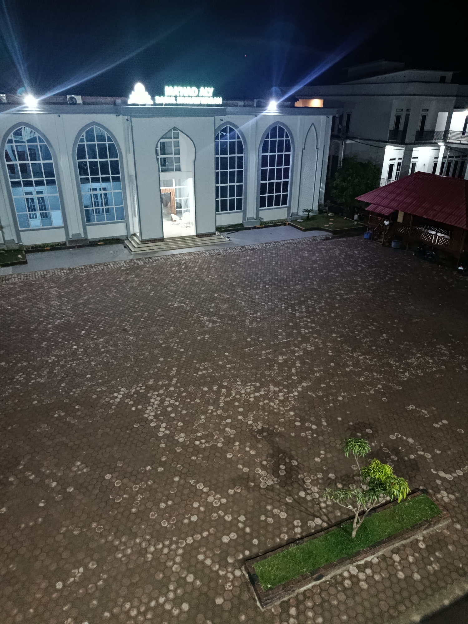 The image depicts an outdoor courtyard at night, possibly a public space or an establishment, located in Peulakancibrek, Indonesia. A modern building with large arched windows and illuminated signage reading "MAJLIS ALIY" and "BANI IBRAHIM IBRAHIM" dominates the left side of the frame. To the right, a rustic gazebo with a red roof is visible, with some seating and people present. The courtyard itself is paved with hexagonal tiles, many of which are wet or have a reflective sheen, suggesting recent rain or moisture. A small, manicured garden bed with a young tree and green grass is situated in the lower right quadrant of the courtyard. The scene is illuminated by artificial lighting from the building and the surrounding environment.