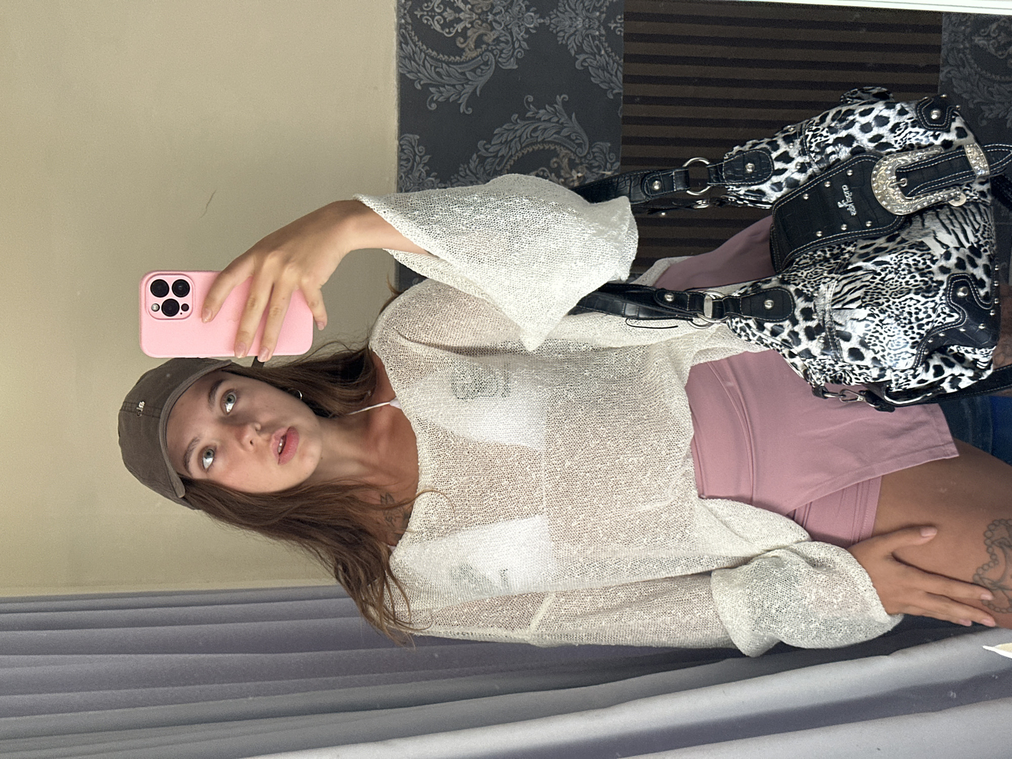 A young Caucasian woman wearing a brown baseball cap, a white sheer top over a white bikini top, and pink shorts stands in front of a mirror, taking a selfie with a pink phone. She holds a black and white leopard-print handbag with silver accents. The background includes a gray curtain and a wall with patterned wallpaper.