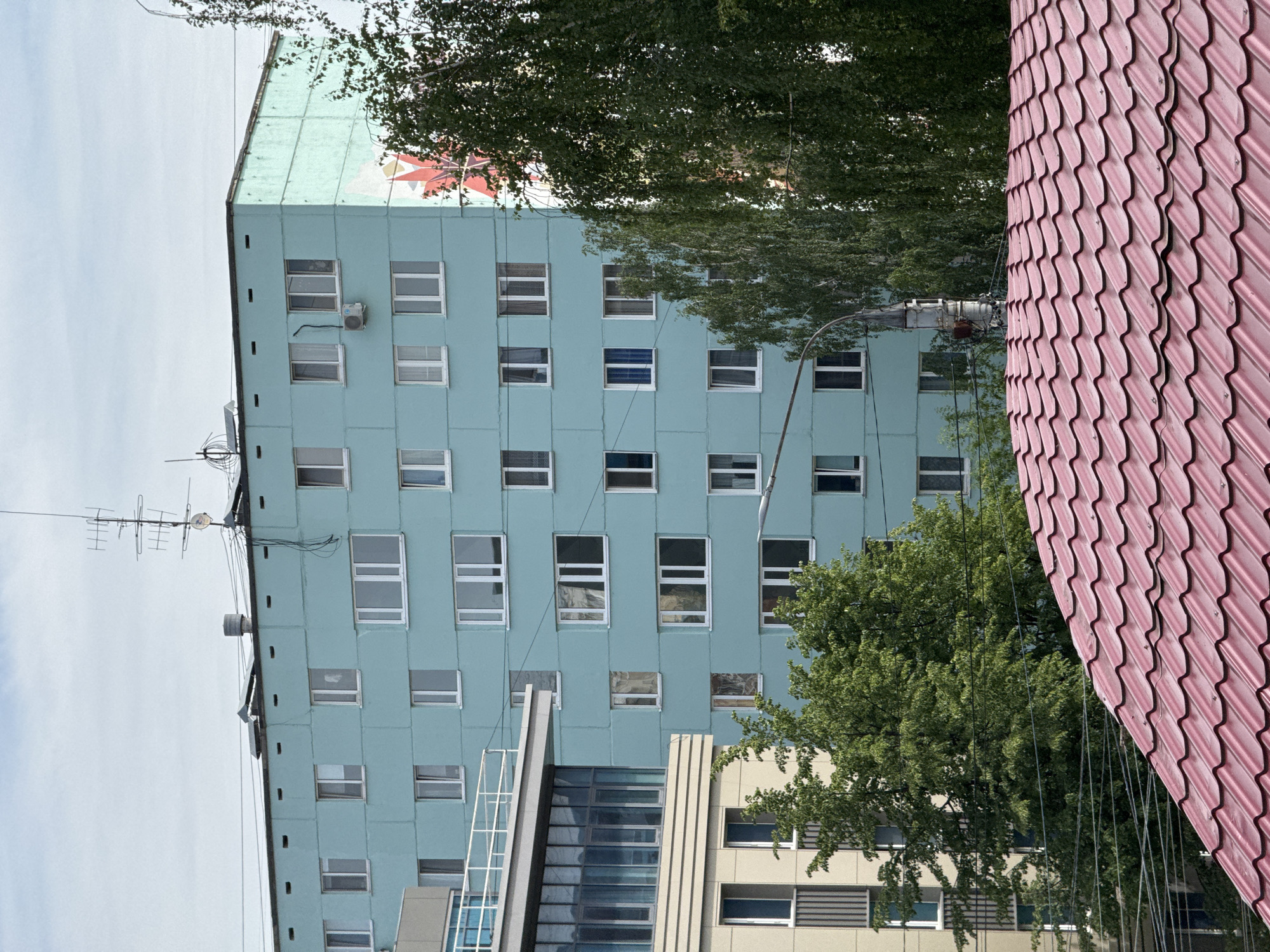 The image captures a multi-story, light-blue building in Izhevsk, Russia. The building features a grid-like facade of numerous windows. At the top of the building, an antenna and a mural can be observed. A street lamp is visible near the building. Trees partially obscure the view of the building and a adjacent structure with a pink roof is in the right corner of the image.