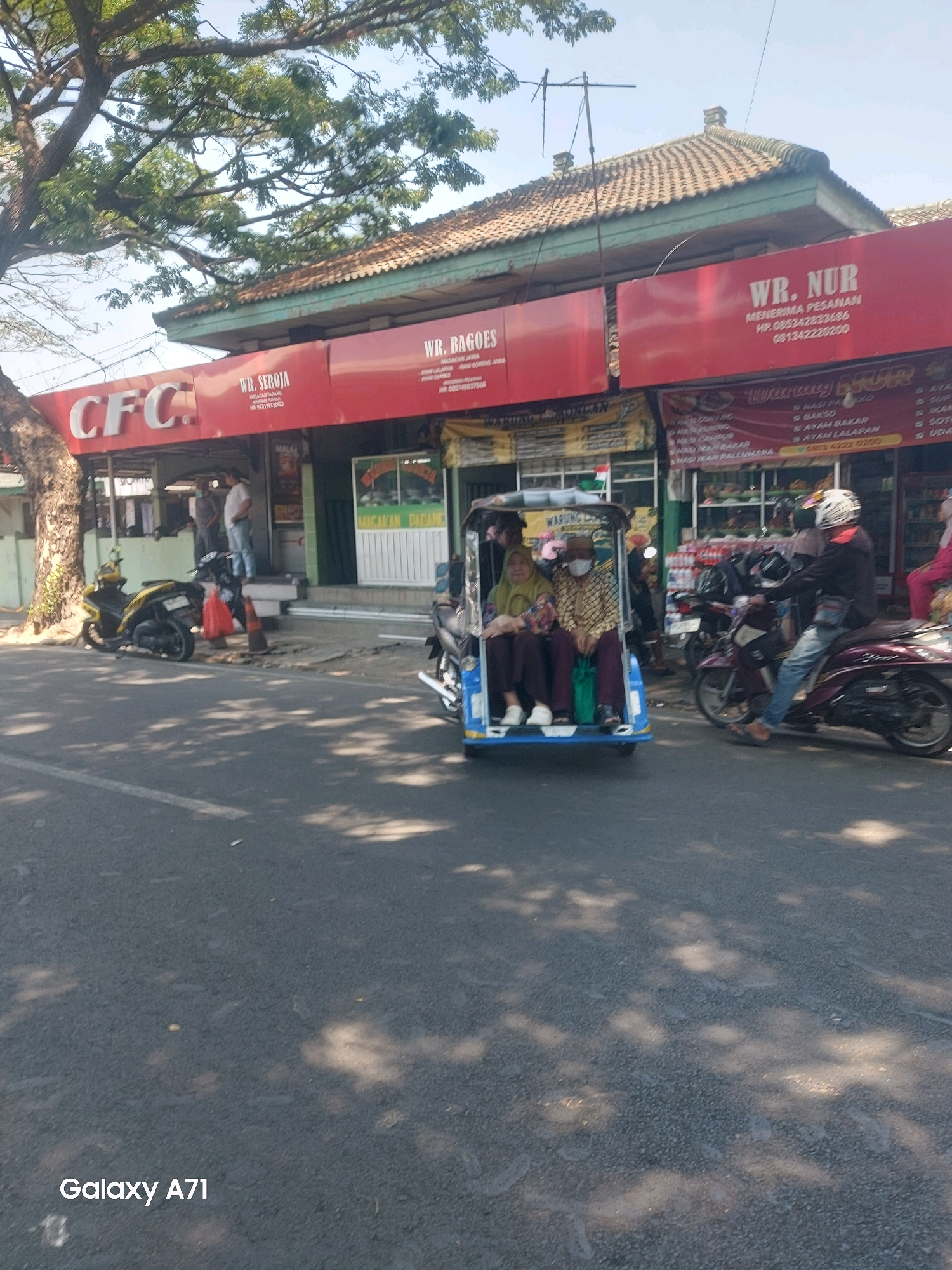The image depicts a street scene in Makassar, Indonesia, on a sunny day, with shadows of trees dappling the asphalt. In the foreground, a road with some white markings is visible. In the middle ground, a blue and white motorized three-wheeled vehicle, often called a "becak motor," is stopped, carrying two women, one wearing a green hijab and a mask, and the other a patterned headscarf and a mask. The becak motor is parked on the side of the road, adjacent to a row of shops.
To the right of the becak motor, a man wearing a helmet and a dark jacket is riding a maroon motorcycle, heading away from the viewer. Further to the right, another person in pink is partially visible. Behind the becak motor and motorcycle, a series of businesses with red signage line the street. One sign reads "CFC." Another sign, also red, displays "WR. SEROJA" with contact information. Adjacent to it, another red sign states "WR. BAGOES." Further down the street, a sign for "WR. NUR" is visible, listing menu items like "NASI GORENG," "NASI PADANG," and "AYAM BAKAR." The shops appear to be open, with some people visible inside or near the entrances. One shop has a green facade with a display of food items behind glass.
The environment suggests a commercial area in an Indonesian city, likely with a mix of food vendors and other small businesses. The presence of the becak motor, motorcycles, and the style of the shops are characteristic of urban settings in Indonesia. The overall impression is a busy street scene during daylight hours.