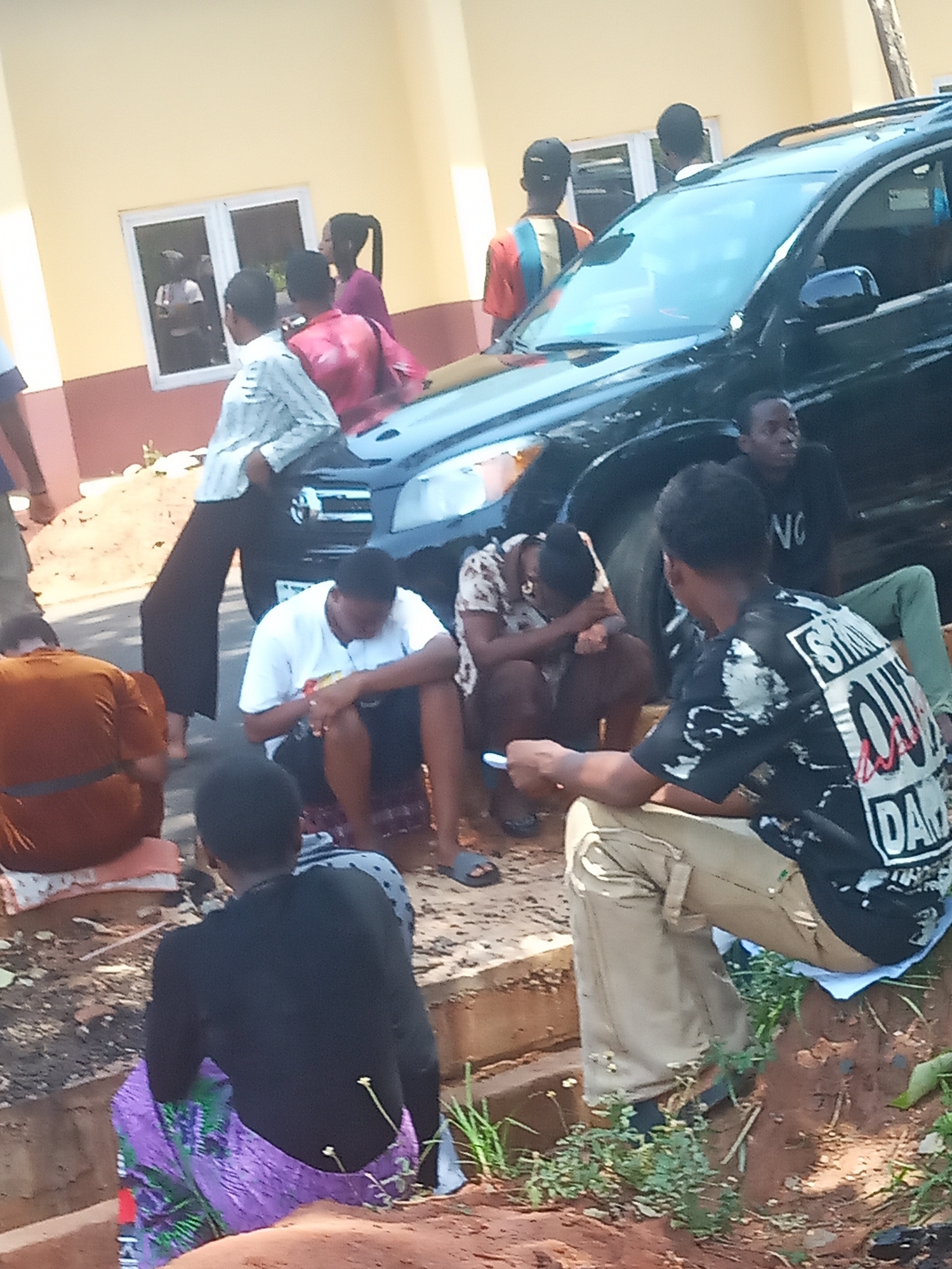 The image depicts a group of people gathered near a black SUV in Ukwu-Abwa, Nigeria. Several individuals are seated on a curb and the ground, some with their heads bowed. Others are standing near the vehicle, and one person is leaning against the car. The setting appears to be outdoors, possibly near a building with a window. The exact actions and interactions within the group are difficult to determine without additional context.
